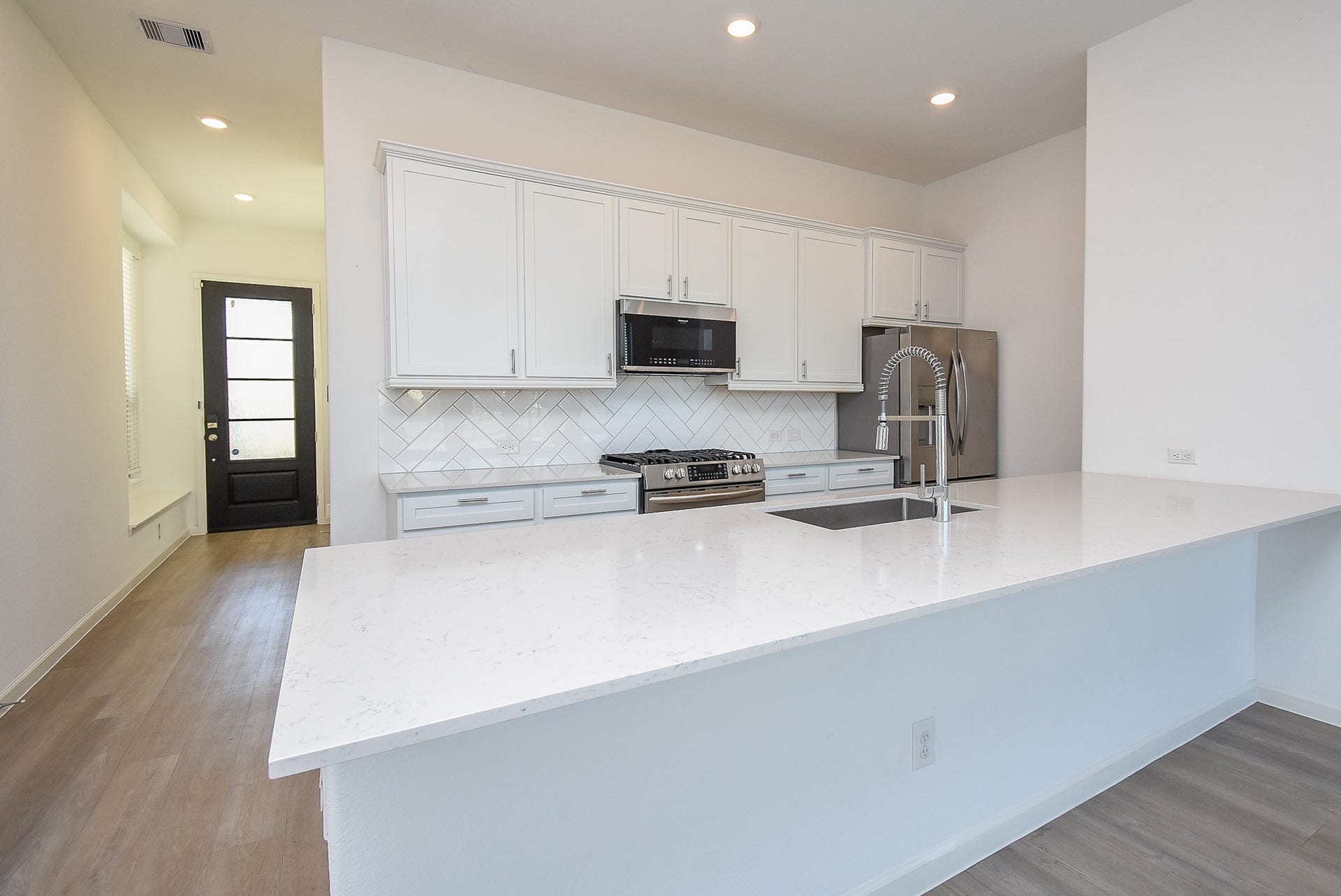 A modern open-concept kitchen with white cabinetry, a large quartz island, and stainless steel appliances. The herringbone tile backsplash adds elegance, while the high-arc pull-down faucet enhances functionality.