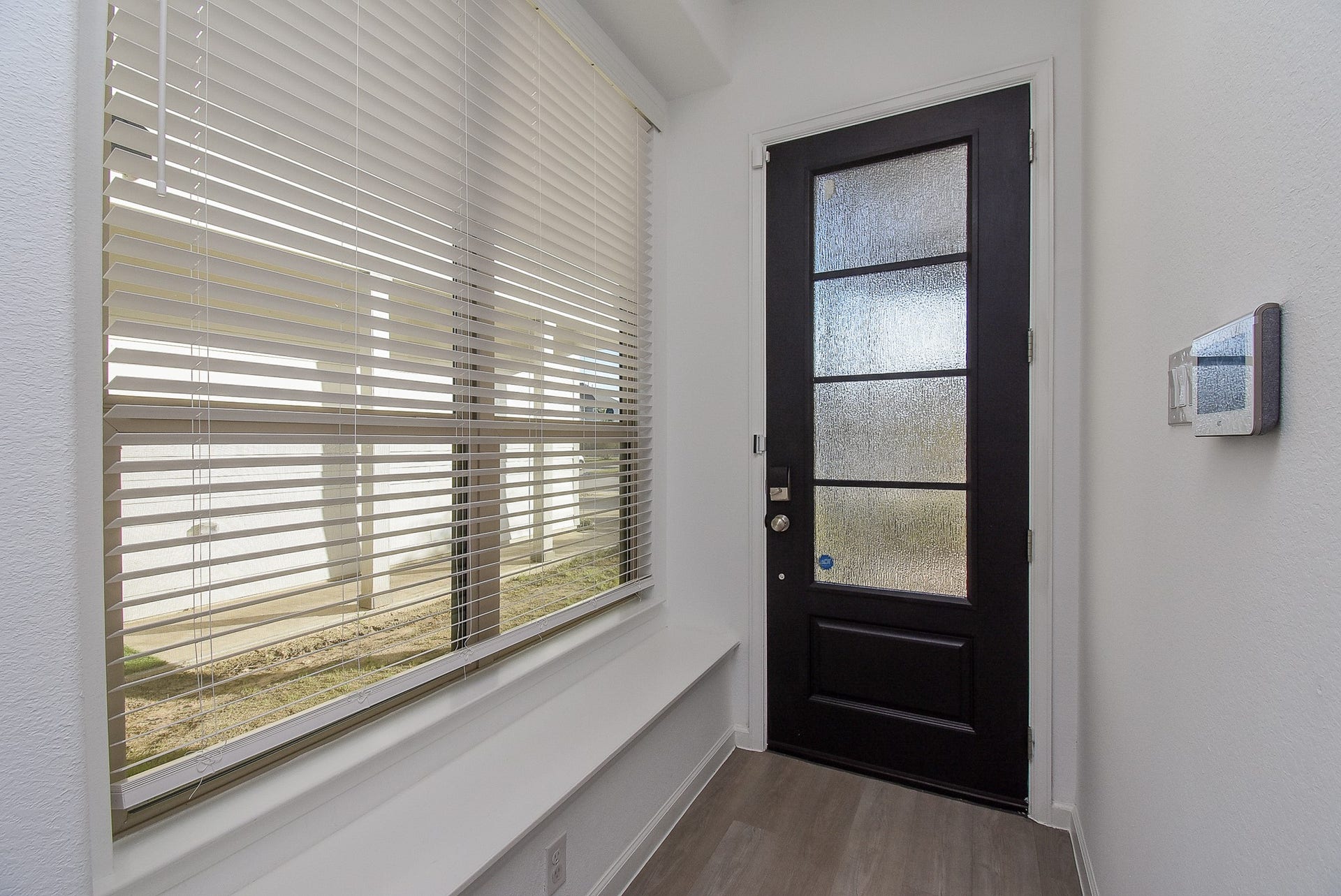 A modern entryway interior featuring a large window with white blinds allowing the natural light to filter in, and a built-in bench runs along the wall