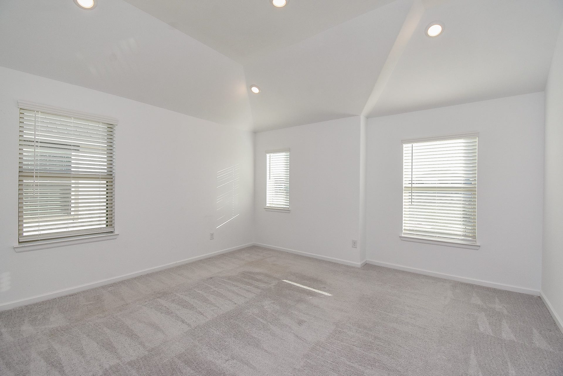 A spacious primary bedroom with neutral tones faces the front of the home, featuring vaulted ceilings with recessed lighting and multiple windows with blinds, allowing for abundant natural light.