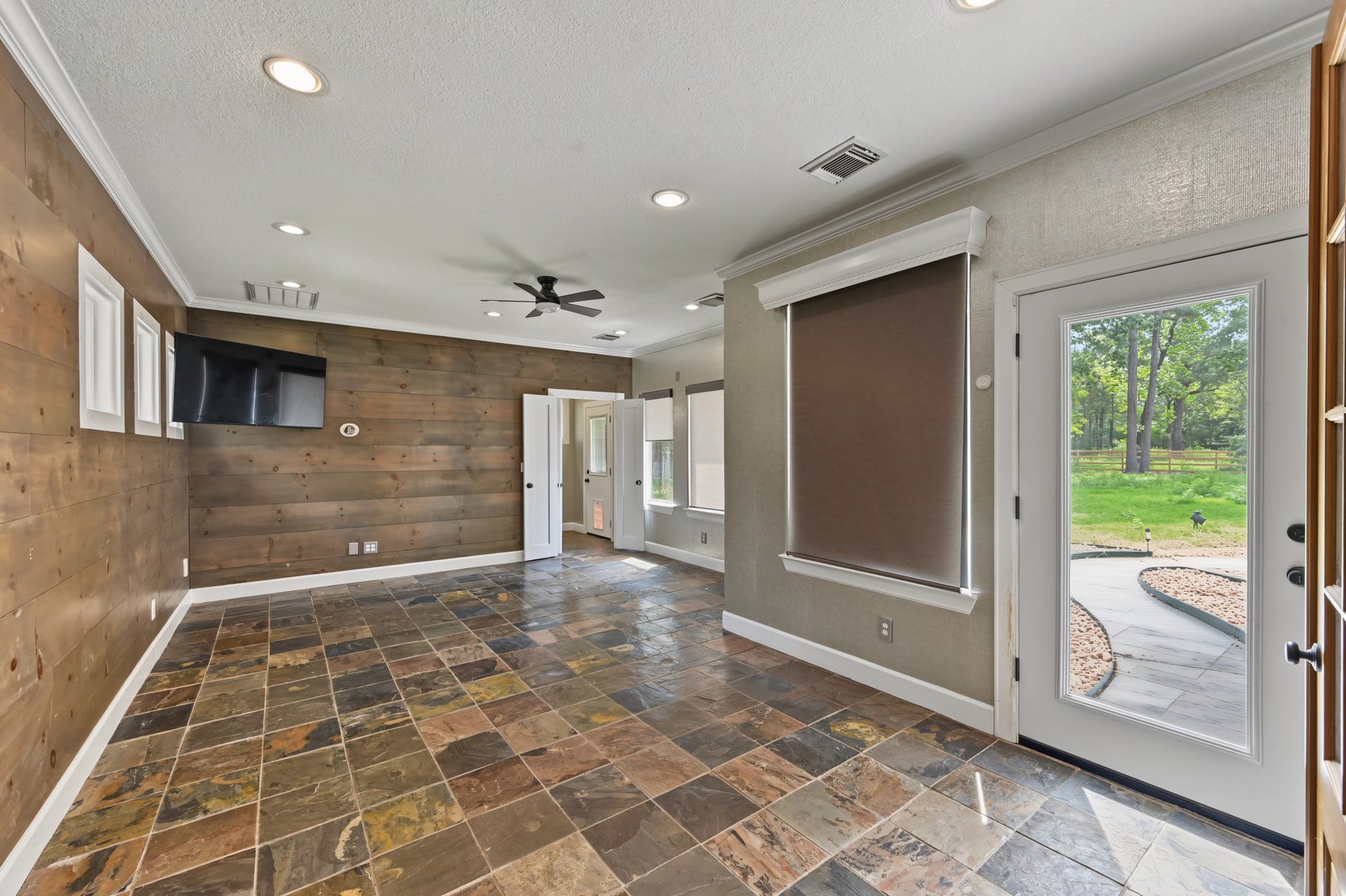 Spacious room used as a gym offers a stylish slate tile floor, rustic wood accent wall, and abundant natural light from large windows and a glass door leading to a serene outdoor view. Also has a full bath that has access just off the backyard/pool area.