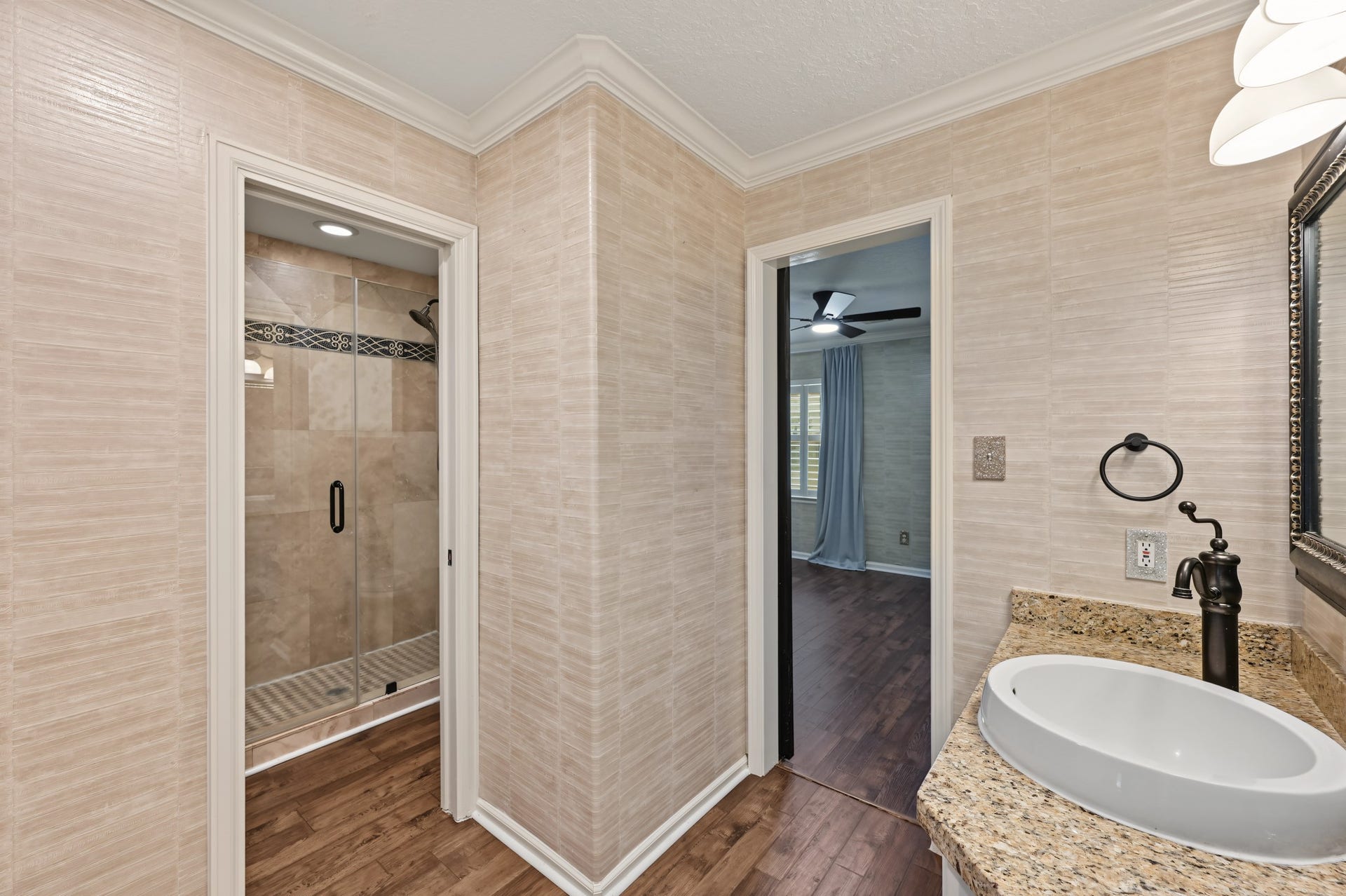 Elegant bathroom with a modern glass-enclosed shower, stylish vessel sink on granite countertop, and warm wood flooring seamlessly leading to the adjacent room.