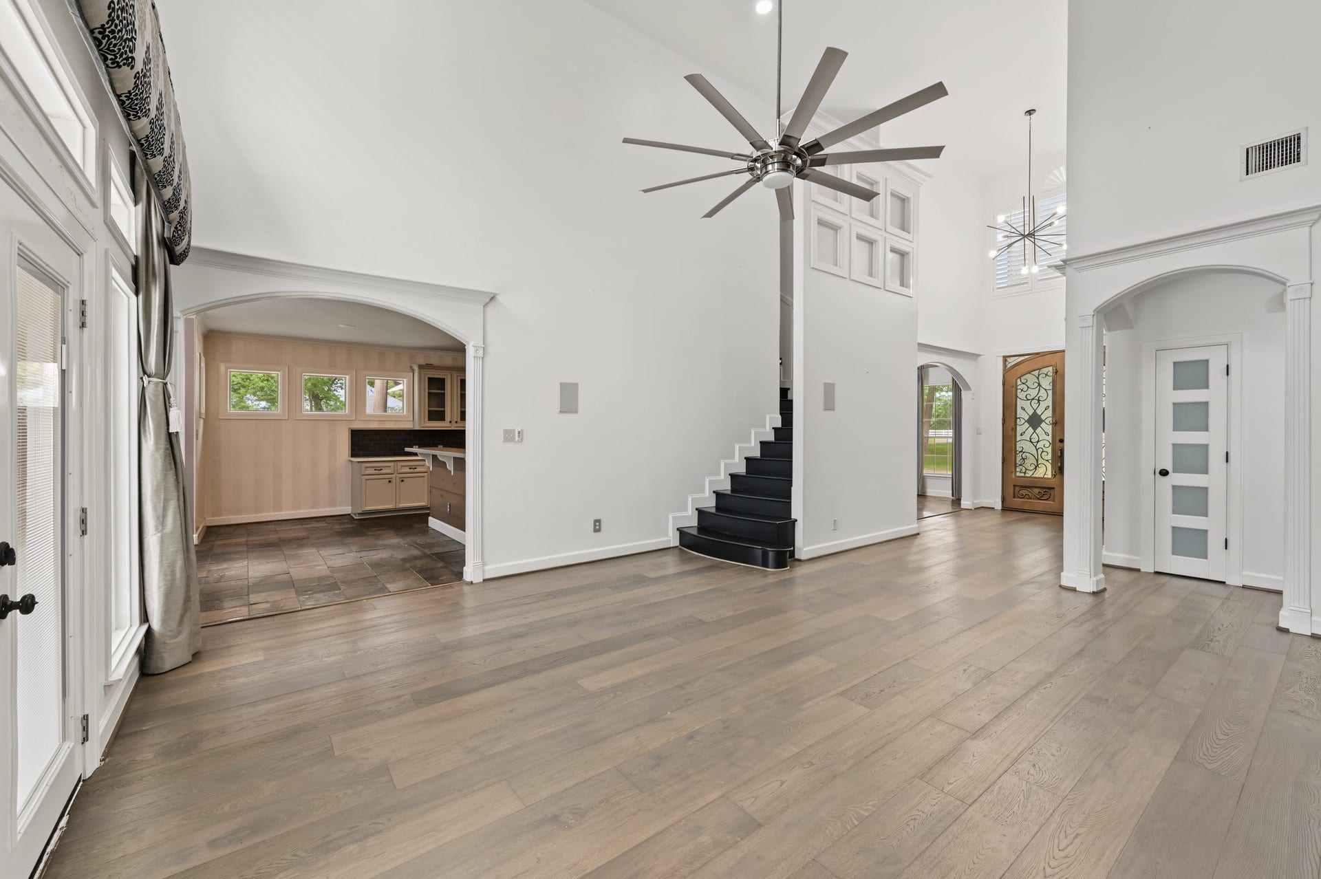 Spacious living area with high ceilings, a striking modern ceiling fan, and elegant wood flooring. The room features an open layout leading to a stylish kitchen and a grand staircase, with abundant natural light and a beautifully detailed front door.