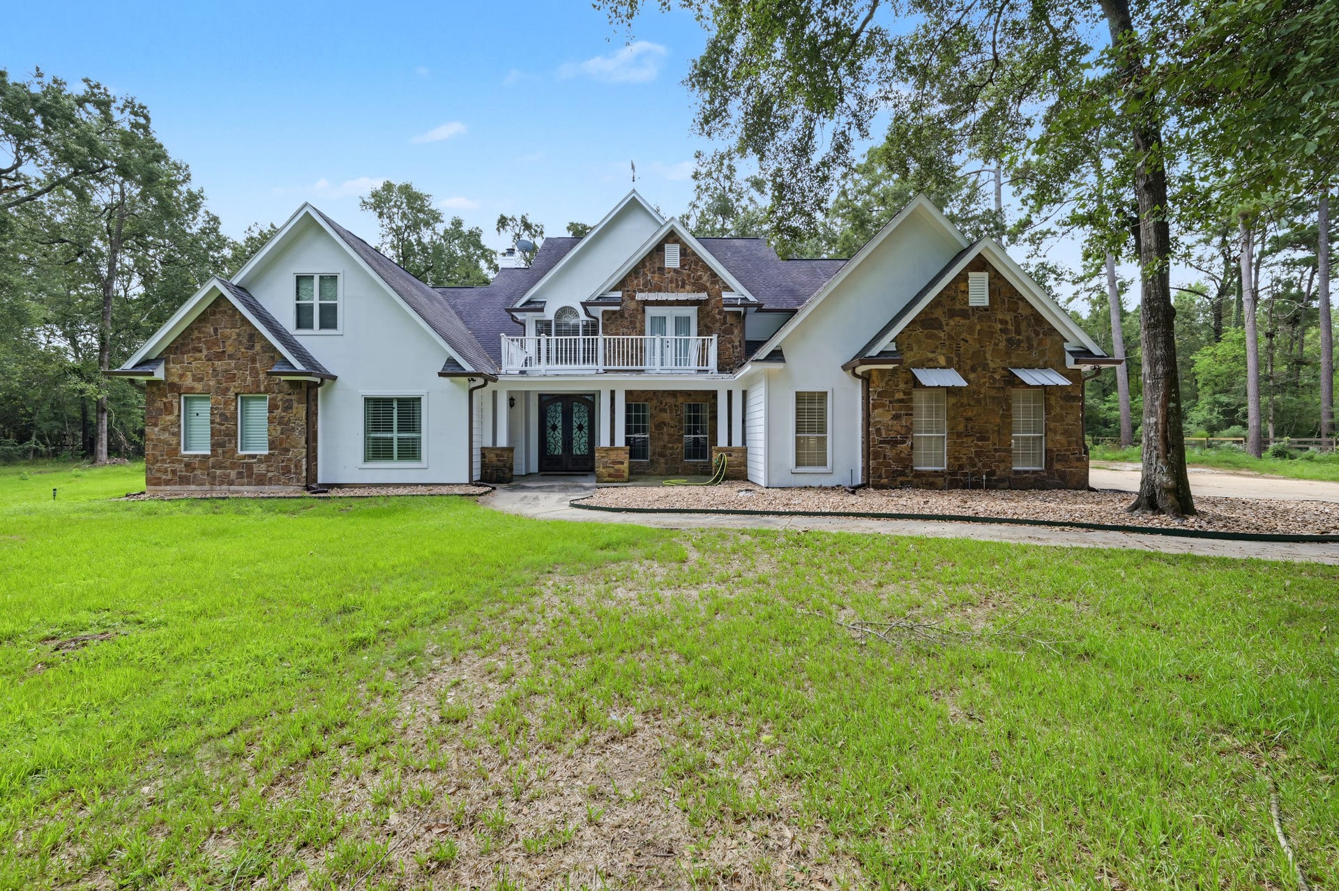 Stunning two-story home with a blend of stone and white siding, nestled in a serene wooded setting. Features a charming front porch, expansive windows, and a spacious lawn.