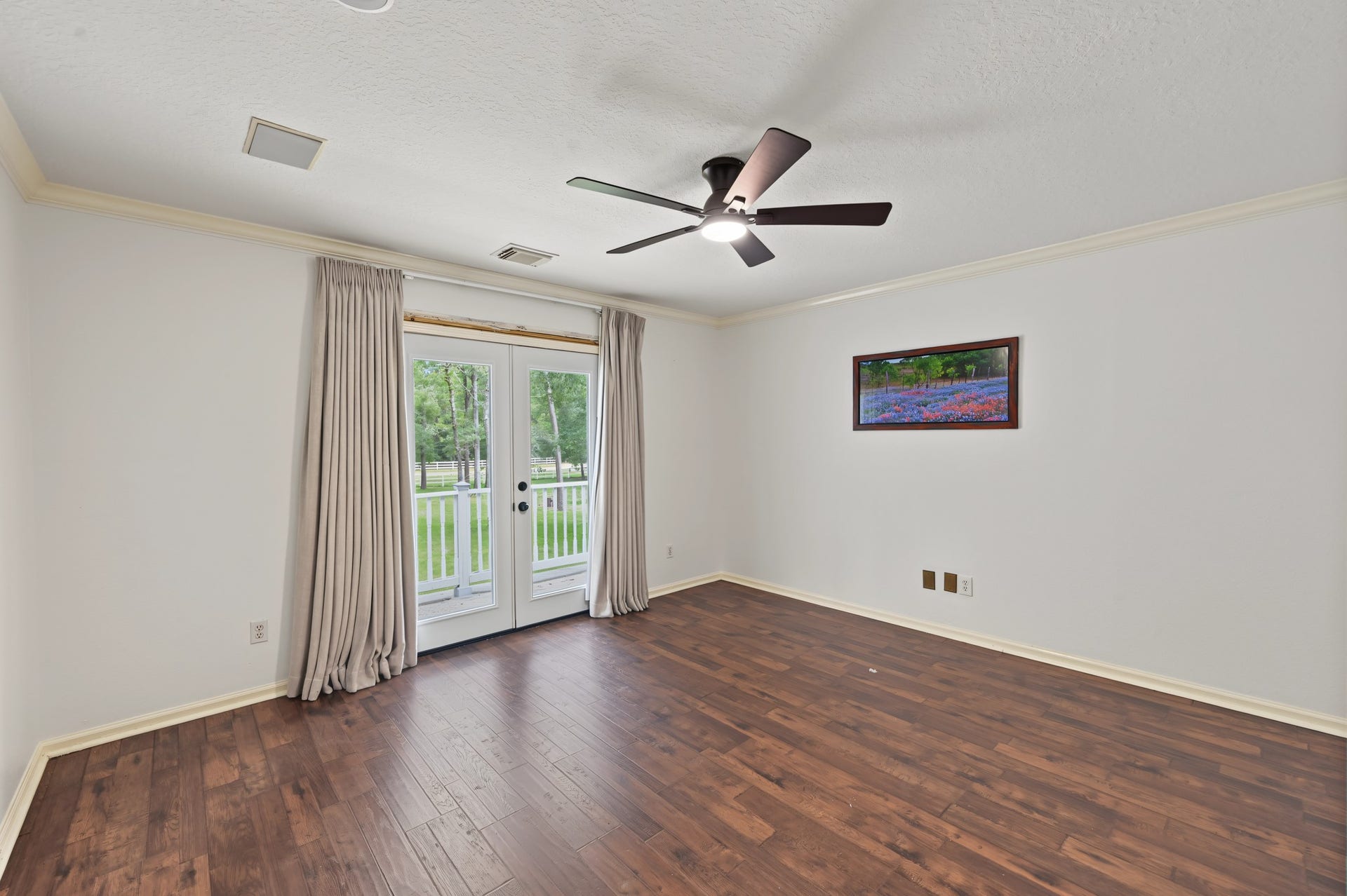 Spacious room with elegant wood flooring, a ceiling fan, and French doors opening to a scenic view, complemented by soft neutral walls and tasteful crown molding.