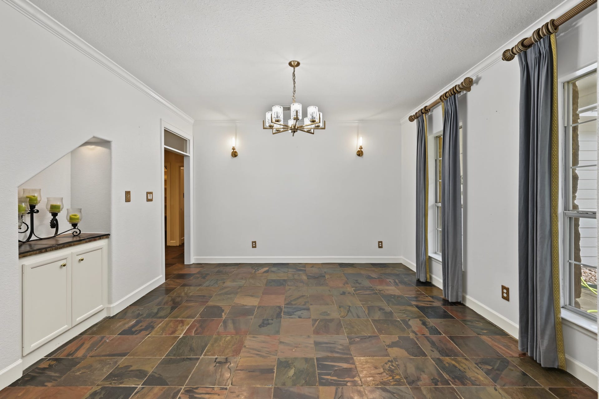 Elegant dining room with slate tile flooring, a stylish chandelier, and large windows with drapes, offering a warm and inviting atmosphere.