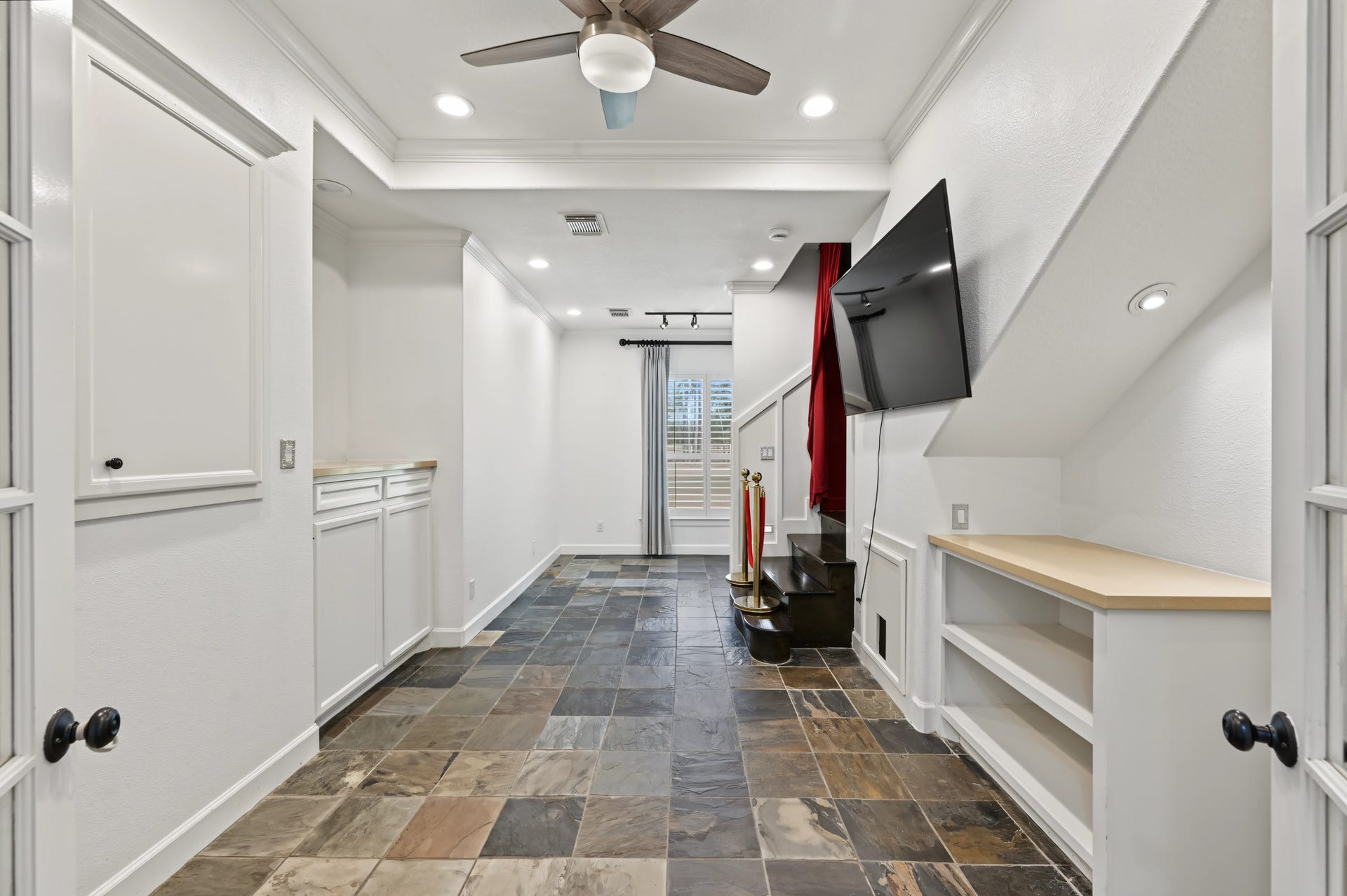 A modern, versatile space featuring slate tile flooring, built-in shelving, and a mounted TV. Recessed lighting and a ceiling fan enhance the bright open feel. Staircase leads to a beautiful theater room!
