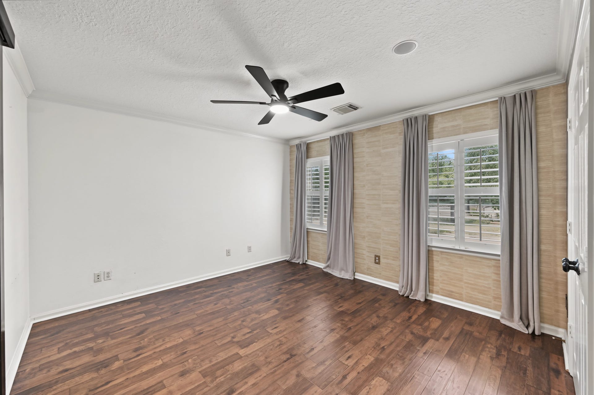Spacious room with rich hardwood floors, abundant natural light from dual windows, elegant curtains, and a modern ceiling fan.