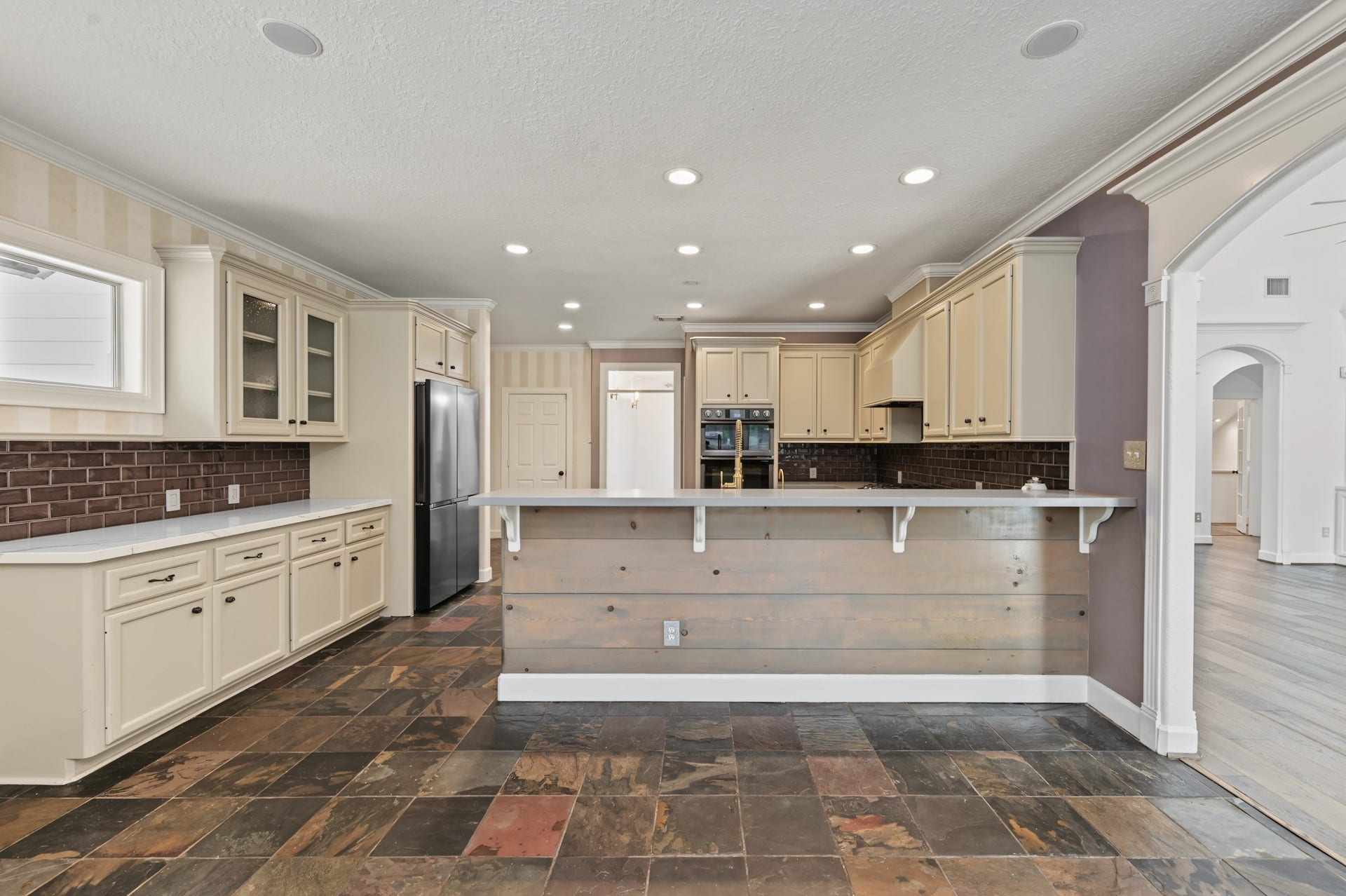 Spacious kitchen with a rustic slate tile floor, elegant cream cabinetry, and a large breakfast bar. The open layout flows seamlessly into the adjoining rooms, complemented by modern stainless steel appliances.