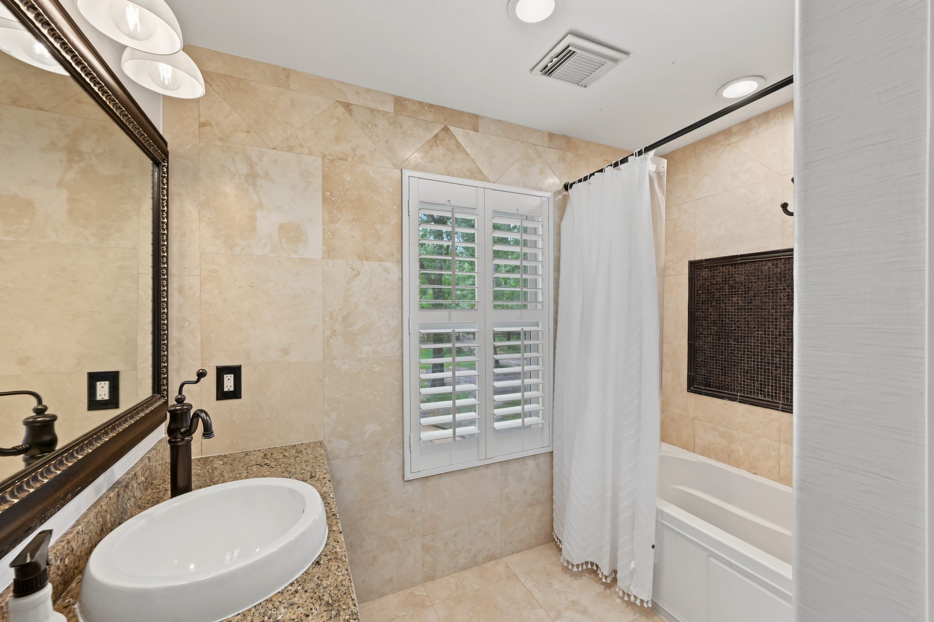Elegant bathroom featuring a granite countertop with a vessel sink, warm tile flooring, and a bright window with plantation shutters.