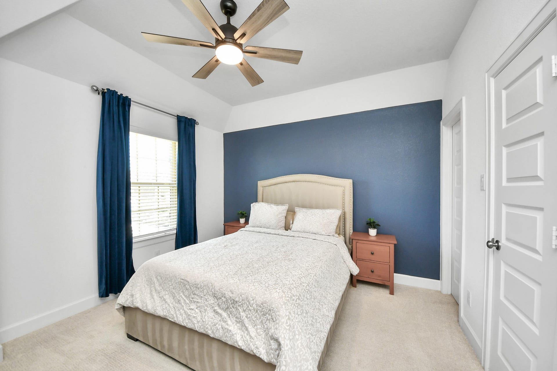 5542 Fairfield Lane, Pasadena, TX 77505 Cozy bedroom featuring a plush bed with a stylish headboard, accented by a bold blue feature wall. Natural light pours in through the window, complemented by elegant blue curtains. A modern ceiling fan enhances the room's comfort.