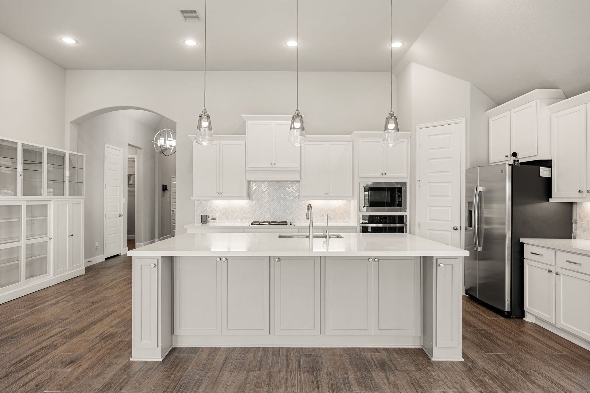 The chefs dream gourmet kitchen serves as the ultimate centerpiece of the home. It features a massive central island topped with pristine engineered quartz, sleek white cabinetry, built-in stainless steel appliances, and a beautiful designer marble backsplash. The space is anchored by three elegant pendant lights and opens beautifully to the main living areas, making it as perfect for meal prep as it is for entertaining.