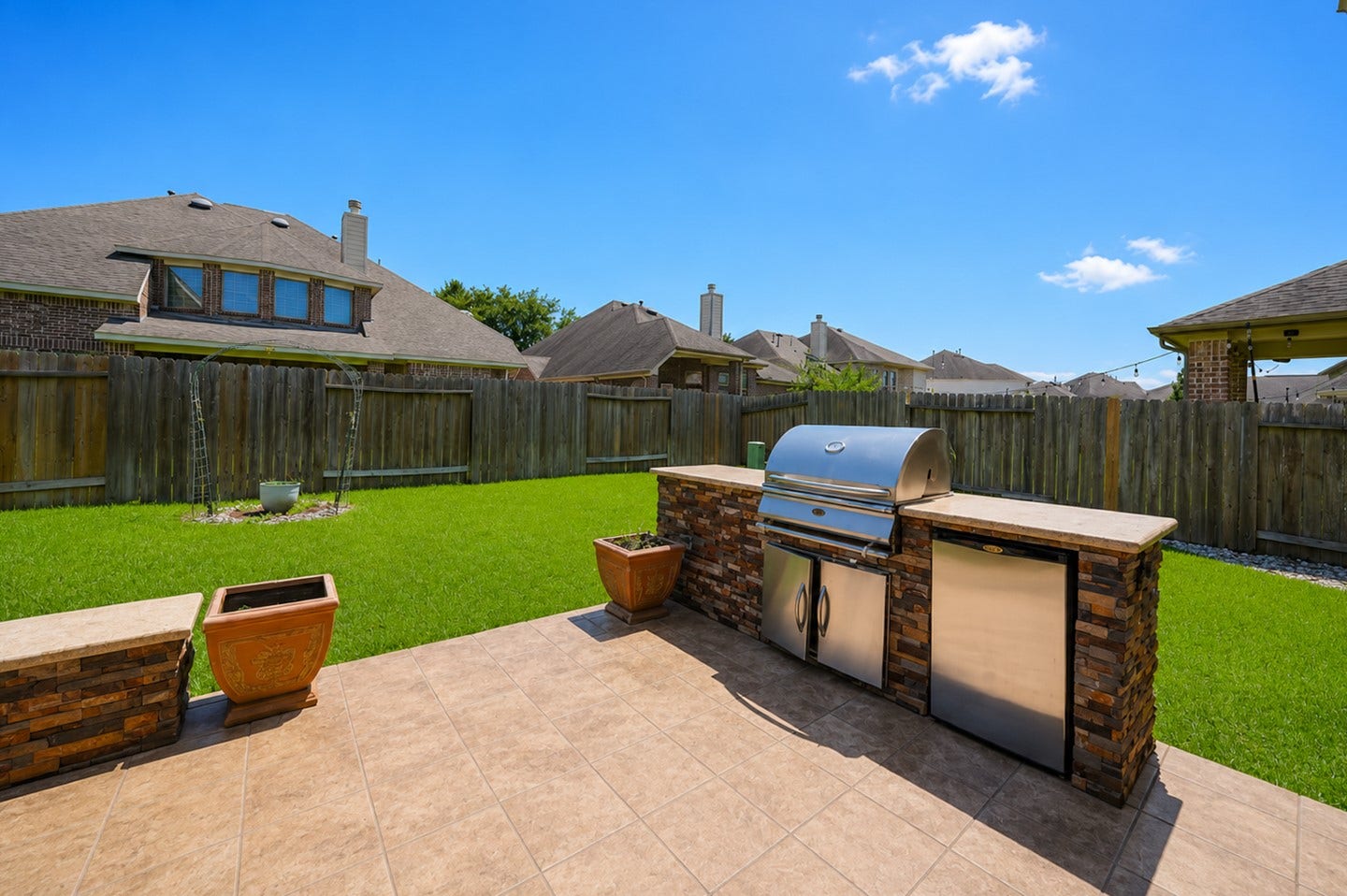 6323 Orange Blossom Lane, Rosenberg, TX 77471 Spacious backyard with a tiled patio featuring an outdoor grill and mini-fridge, perfect for entertaining. Well-maintained lawn and privacy fence enhance the outdoor living space.