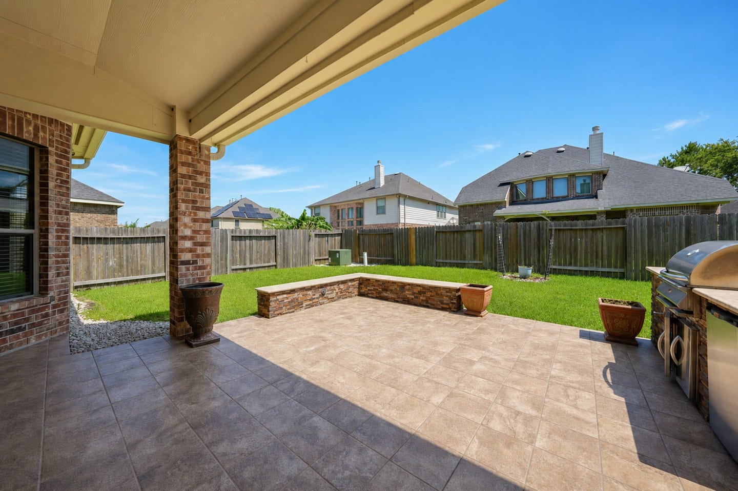 6323 Orange Blossom Lane, Rosenberg, TX 77471 This photo showcases a spacious, covered patio with tiled flooring, perfect for outdoor entertainment. It includes a built-in grill and overlooks a well-maintained, fenced backyard with a small stone bench, providing privacy and a great space for relaxation.