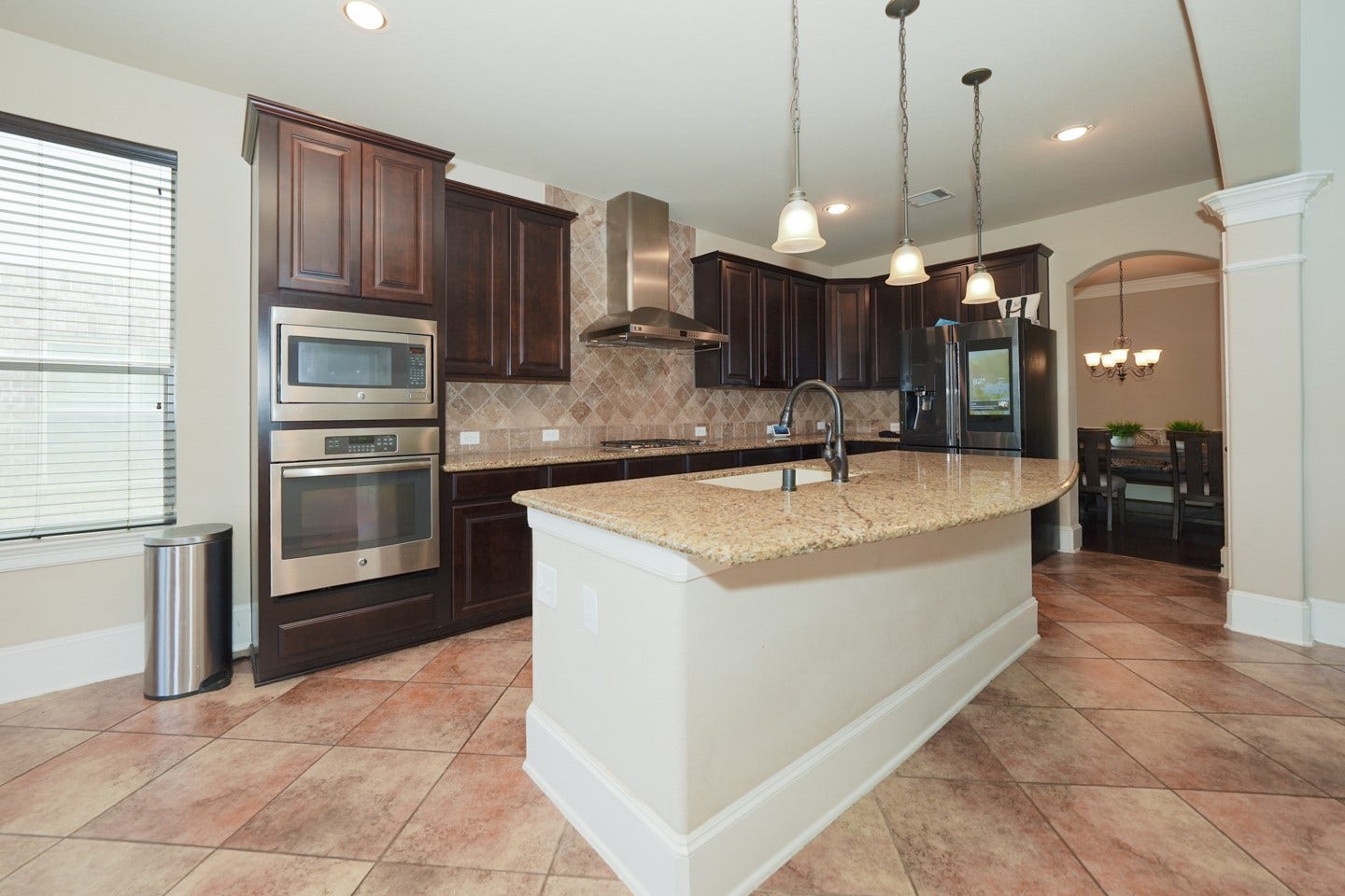 6323 Orange Blossom Lane, Rosenberg, TX 77471 This modern kitchen features dark wood cabinets, stainless steel appliances, and a large granite-topped island with pendant lighting. The open layout flows into a dining area, highlighted by tiled floors and ample natural light.