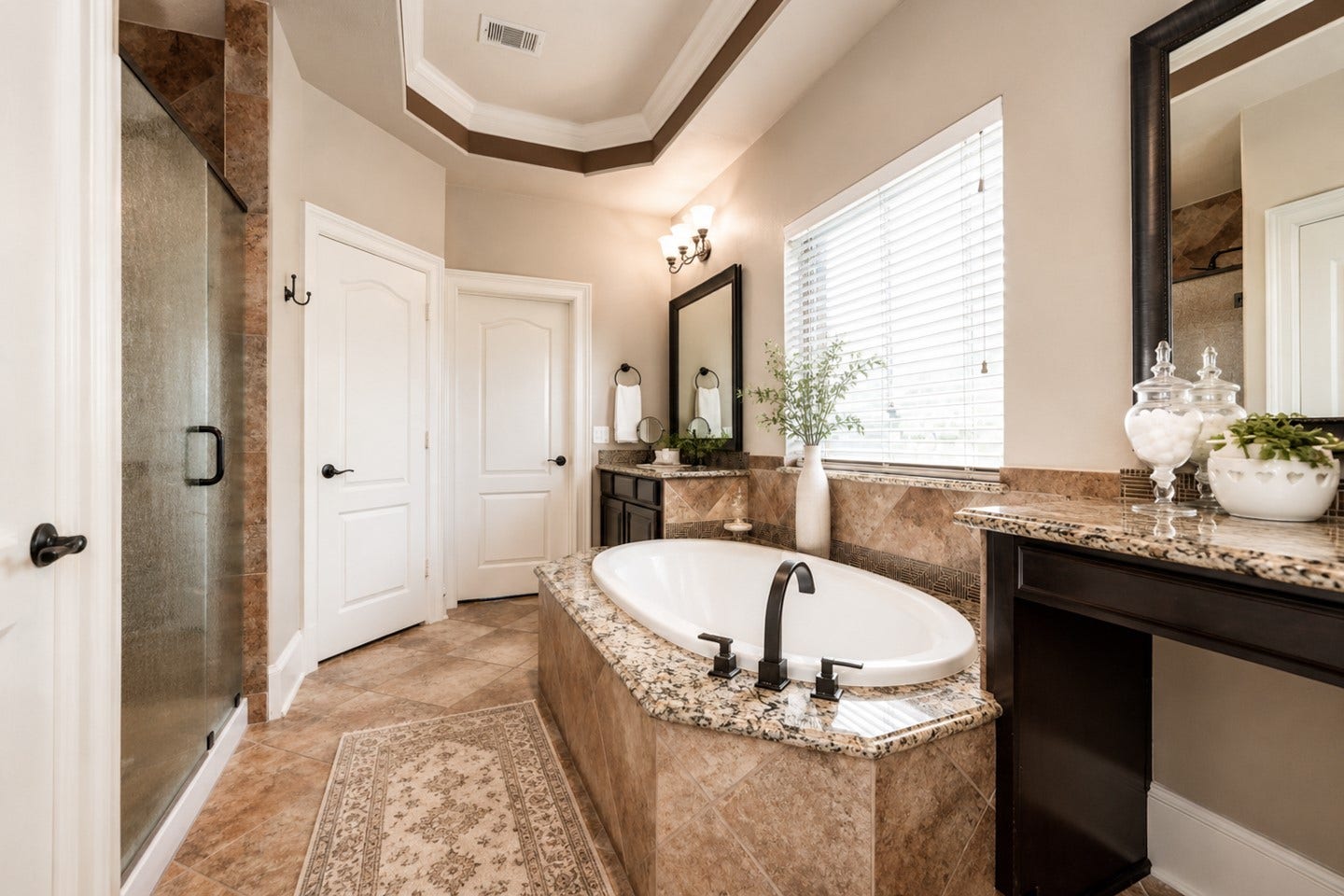 6323 Orange Blossom Lane, Rosenberg, TX 77471 This elegant primary bathroom features a central soaking tub with granite surround, a spacious glass-enclosed shower, dual vanities with large mirrors, and tasteful tile flooring. Natural light filters through a window with blinds, creating a bright and inviting space.