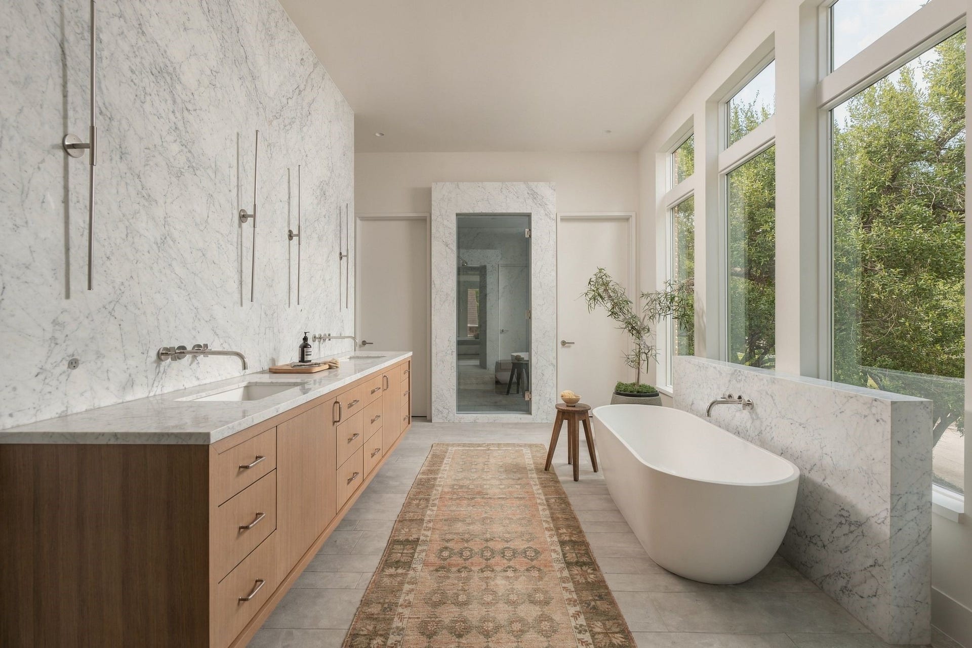 A breathtaking Venatino marble-clad primary bath designed as a true retreat. Large floating rift white oak vanity w/dual sinks, wall-mounted Watermark fixtures & sleek Juniper lighting is complemented by a pedestal soaking tub & incredible walled-in shower finished in marble, limestone & concrete.