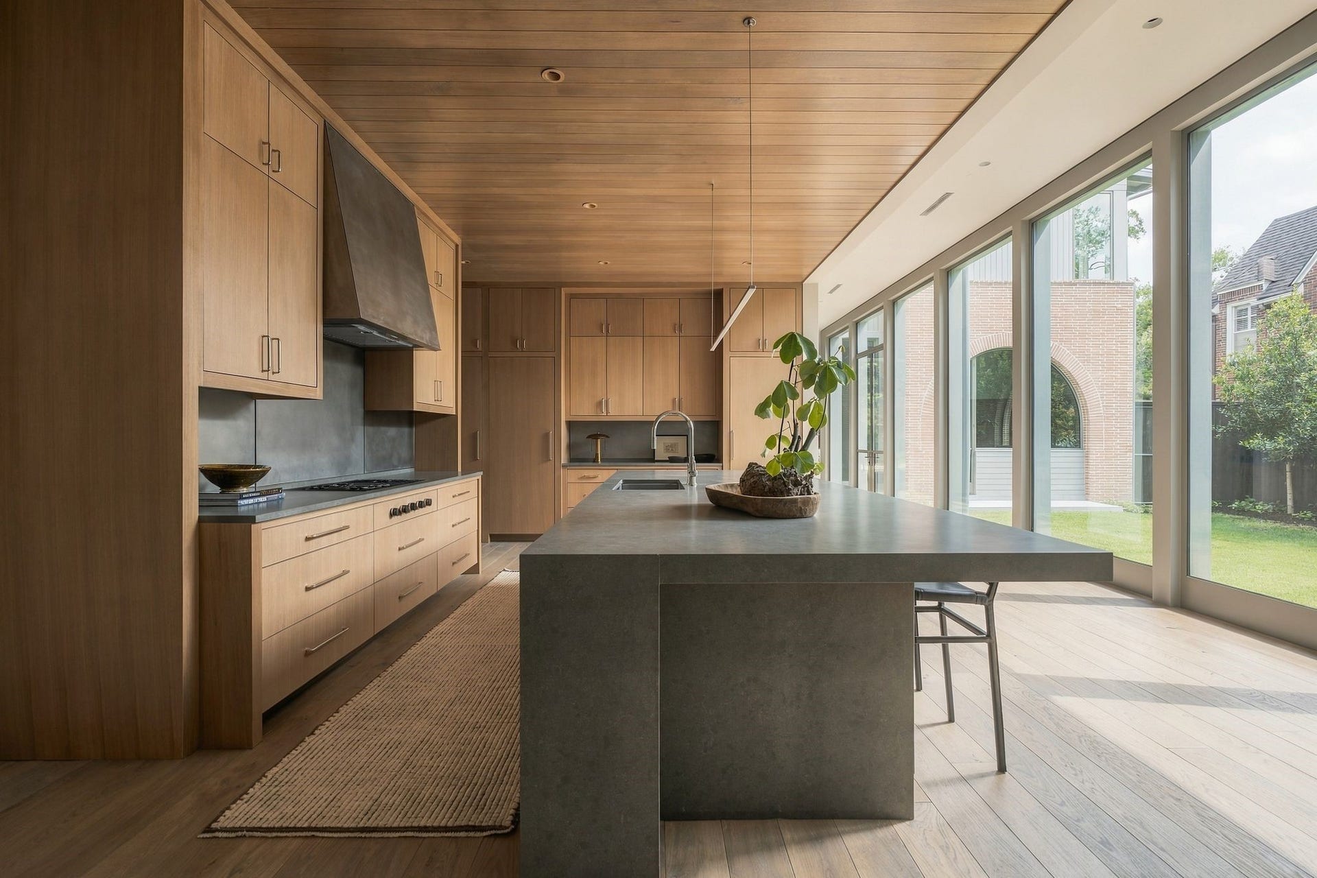 From the family room, the craftsmanship of the kitchen comes fully into viewrift white oak cabinetry, white oak-paneled ceiling, custom stainless detailing & premium appliances. A continuous wall of windows runs alongside both spaces, filling the interiors w/natural light & greenery.