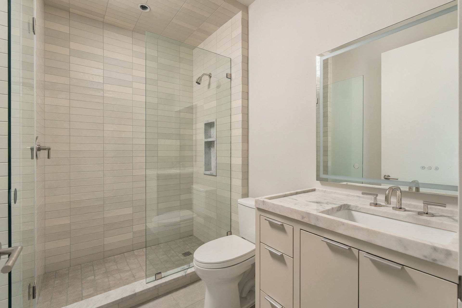 Another private en suite guest bath features a walk-in shower w/porcelain subway tile surround, built-in soap niche & frameless glass. Limestone flooring, generous vanity storage, polished nickel fixtures & an illuminated inset mirror create a clean, timeless finish.