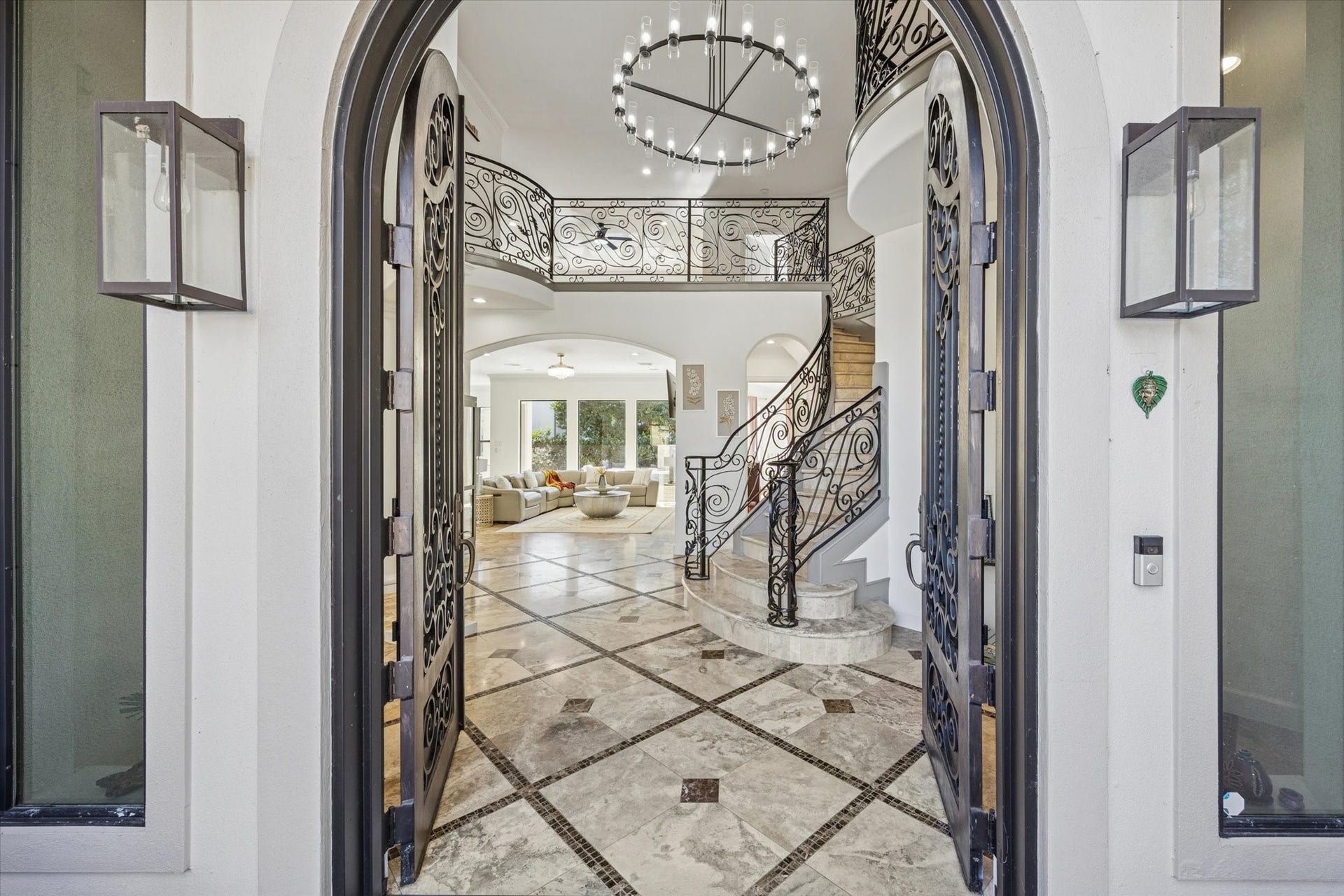 61 Open Sands Court, Sugar Land, TX 77479 This elegant entryway features double iron doors opening to a grand foyer with a curved staircase and intricate wrought iron railings. The space is bright and open, with a large chandelier and travertine and marble inlay flooring, leading into a spacious living area with ample natural light.