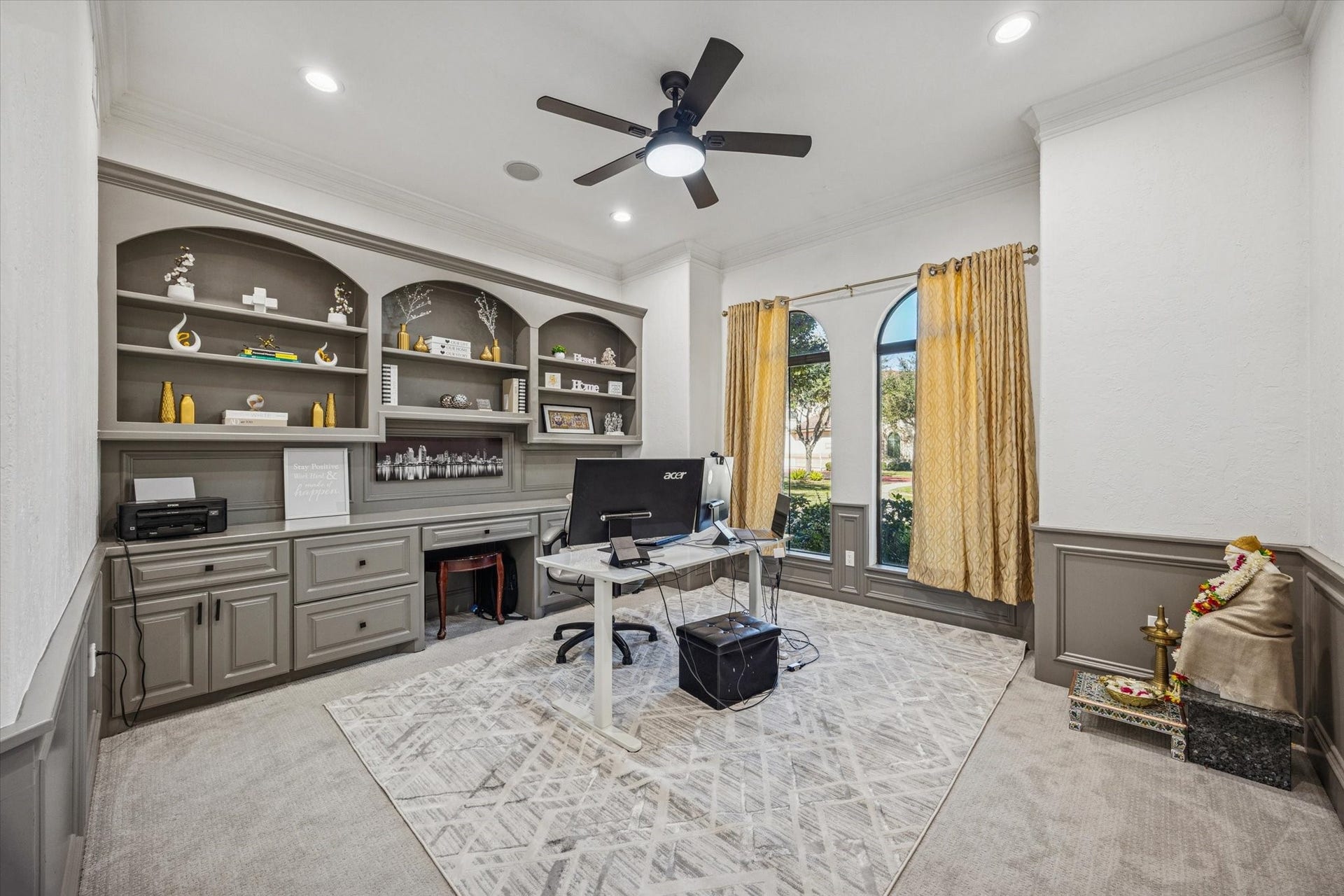 61 Open Sands Court, Sugar Land, TX 77479 This photo showcases a bright home office with built-in shelves and cabinets, a ceiling fan, large windows with drapes, and neutral-colored walls and carpet. Ideal for a functional workspace.