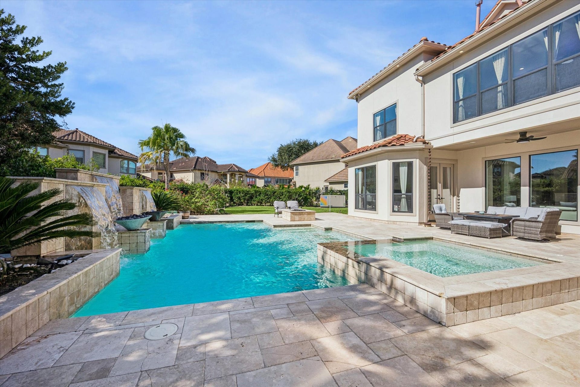 61 Open Sands Court, Sugar Land, TX 77479 This photo showcases a luxurious outdoor space with a sparkling pool and spa, surrounded by elegant stone tiles. The modern two-story home features large windows and a spacious patio area with comfortable seating, perfect for relaxing or entertaining.