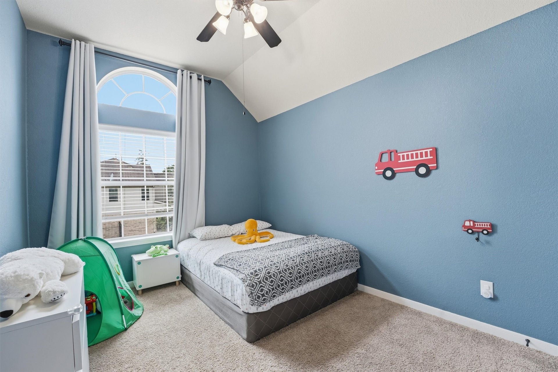 Bright, neutral-toned bedroom with soft carpeting, a large window, and a ceiling fan. Ideal for comfort and relaxation.