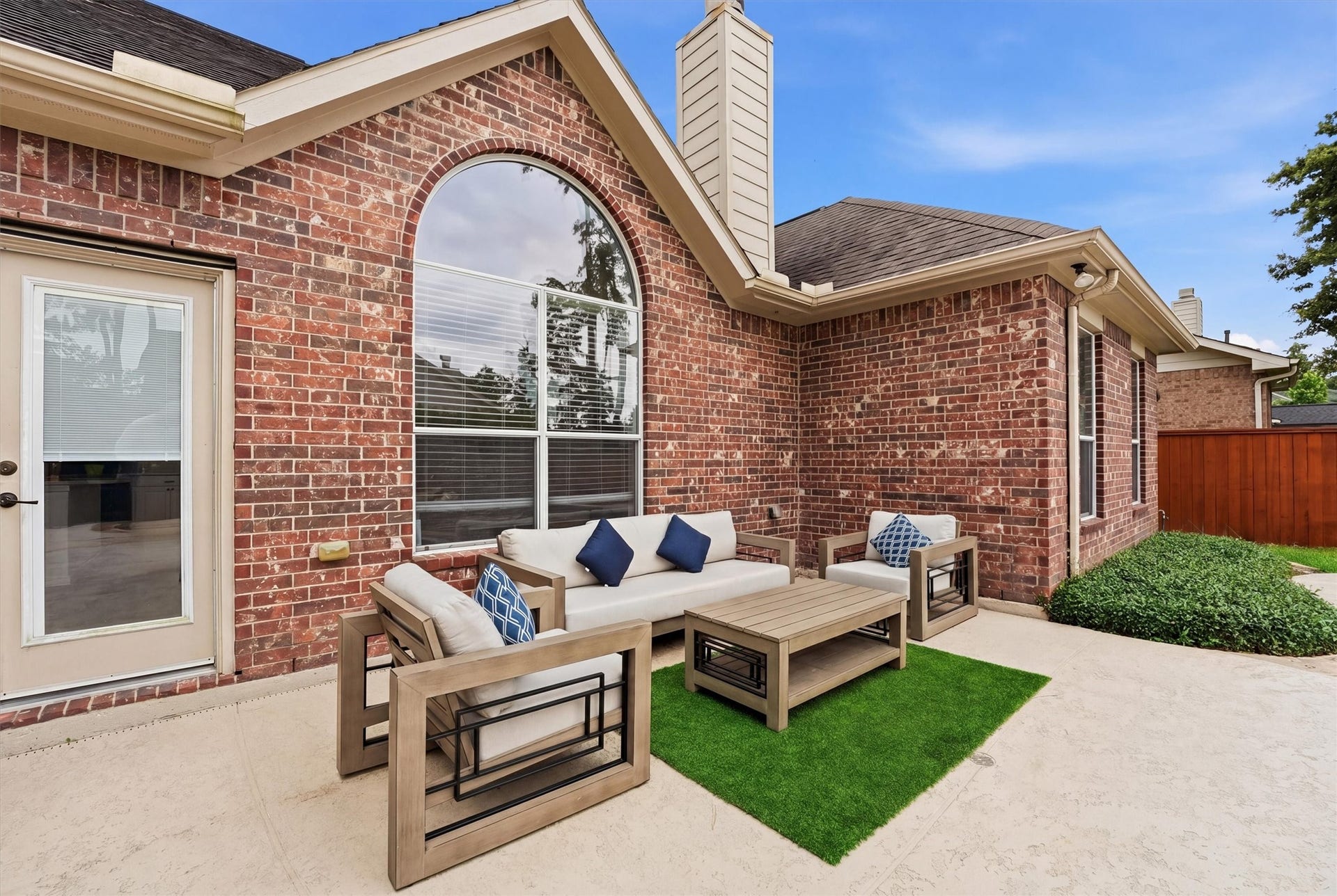 This photo showcases a charming outdoor patio area with a brick exterior, featuring a cozy seating arrangement with a sofa and armchairs around a coffee table on a small green rug. The space is well-lit with a large arched window and a nearby glass door, providing a welcoming atmosphere for relaxation or entertaining.