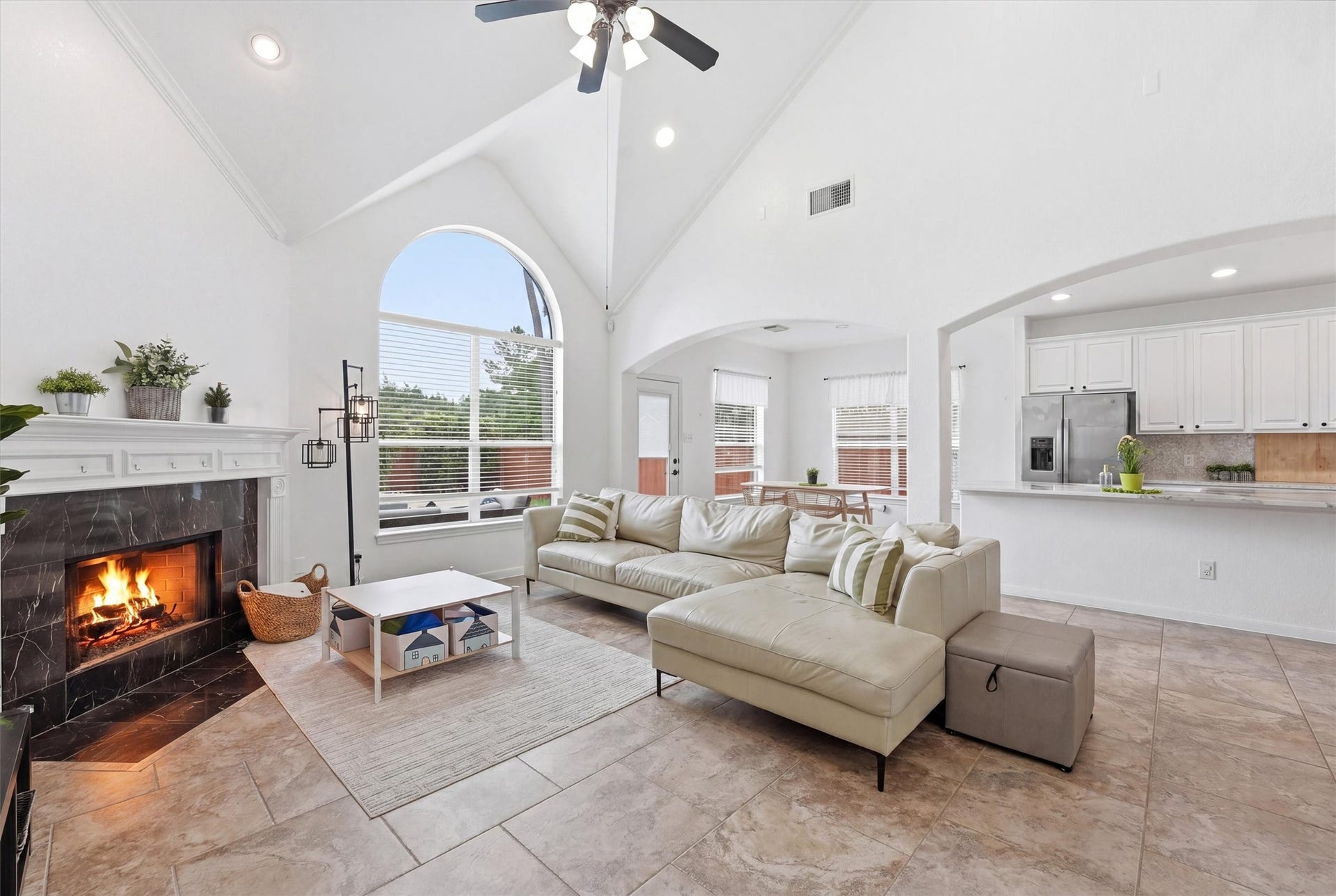 This spacious living room features high vaulted ceilings, a cozy fireplace, and a large arched window for abundant natural light. It's complemented by a modern ceiling fan, tile flooring, and an open, inviting layout.