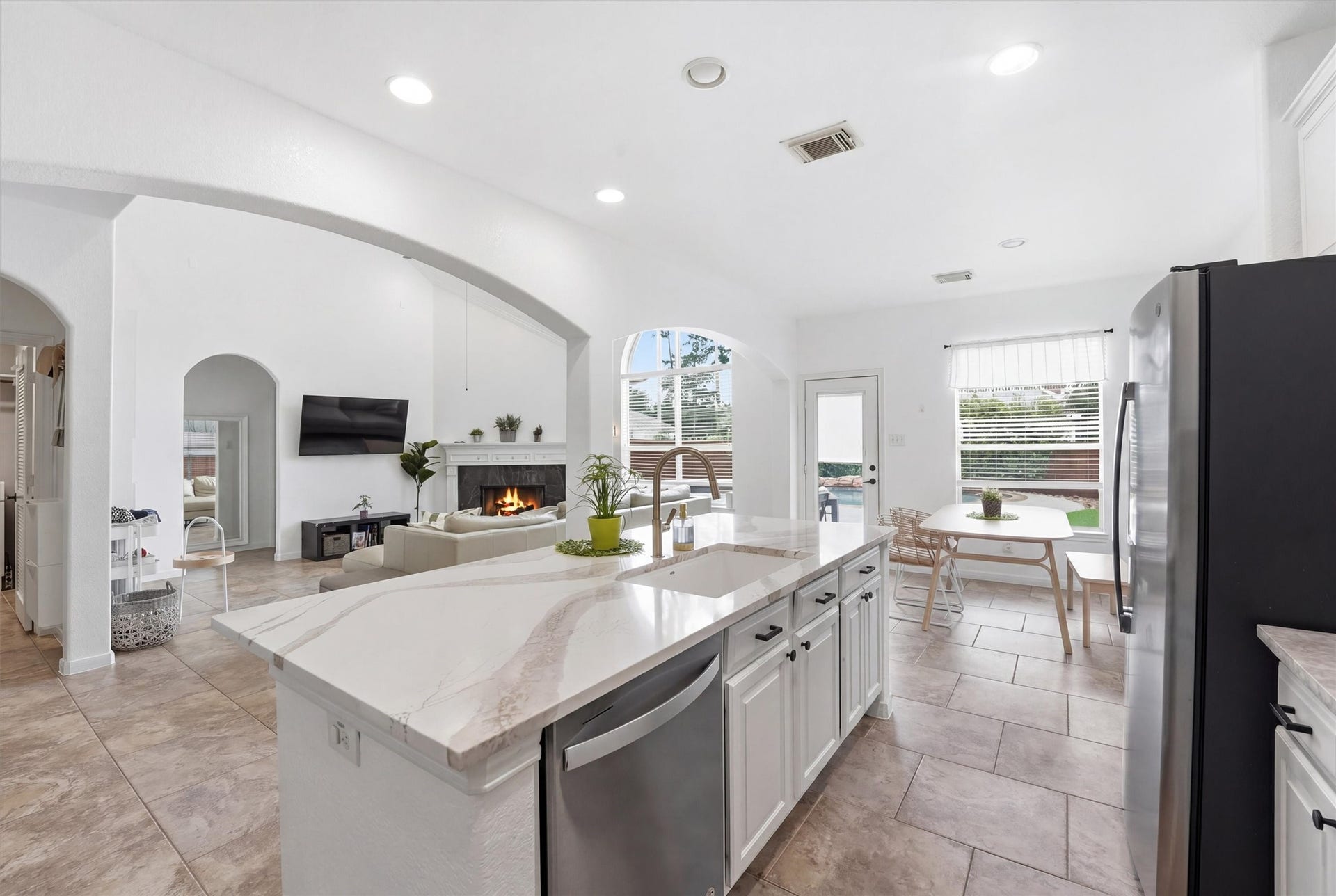 This modern kitchen features sleek stainless steel appliances, ample white cabinetry, a spacious marble-topped island, and elegant tile flooring. Arched doorways add charm, leading to an open living area, creating a seamless flow perfect for entertaining.
