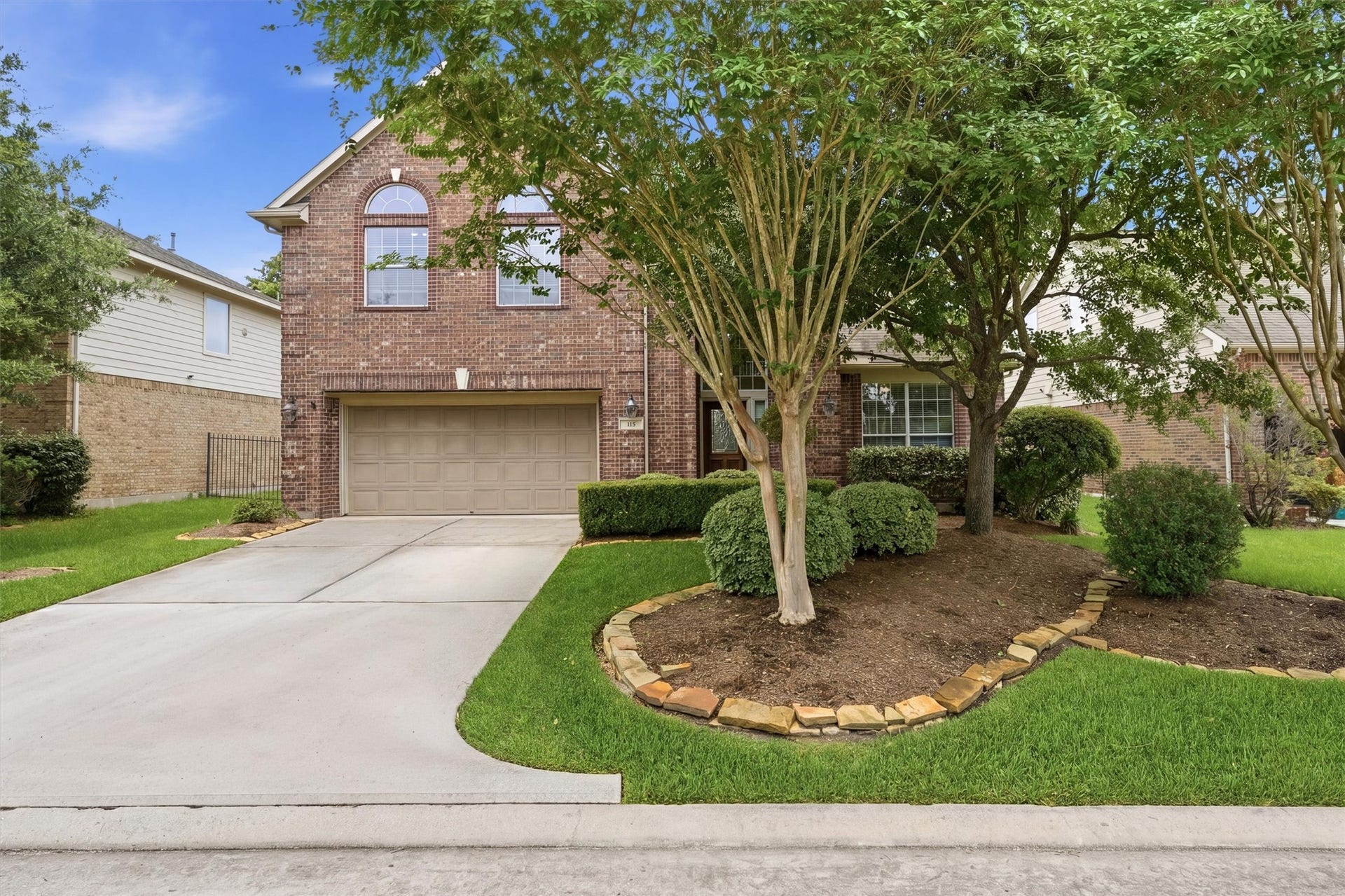 This photo shows a two-story brick house with a two-car garage and a well-maintained front yard. The yard features mature trees and neatly trimmed shrubs, enhancing the curb appeal. The driveway is spacious, and the neighborhood appears inviting and well-kept.