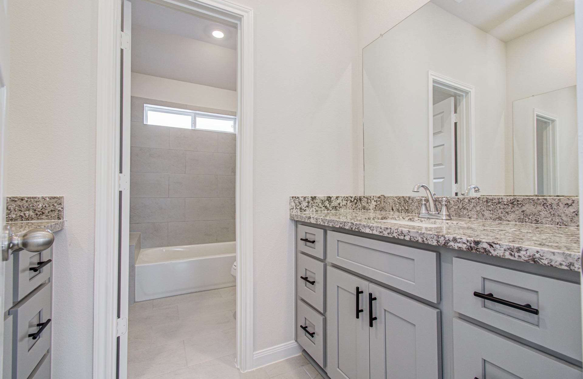 14210 Aston Pine Drive, Houston, TX 77032 Elegant bathroom featuring a spacious granite-topped vanity with sleek gray cabinetry, leading to a bright tiled tub area with natural light from a high window. Please Note this is a different home/floorplan in the community.