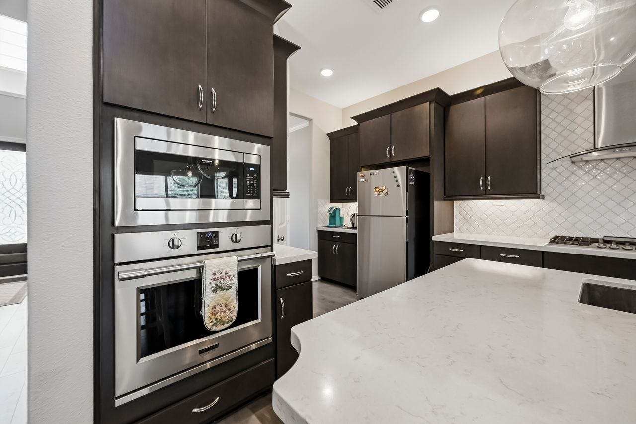 2215 Hermina Radler Drive, Richmond, TX 77469 This sophisticated kitchen centers on high-end functionality, showcasing built-in stainless steel ovens set against rich, dark cabinetry. A sleek professional range hood anchors the space, while expansive light-toned countertops with delicate grey veining offer ample room for preparation. Tucked beyond the main workspace, a dedicated breakfast and coffee nook completes this refined culinary environment.