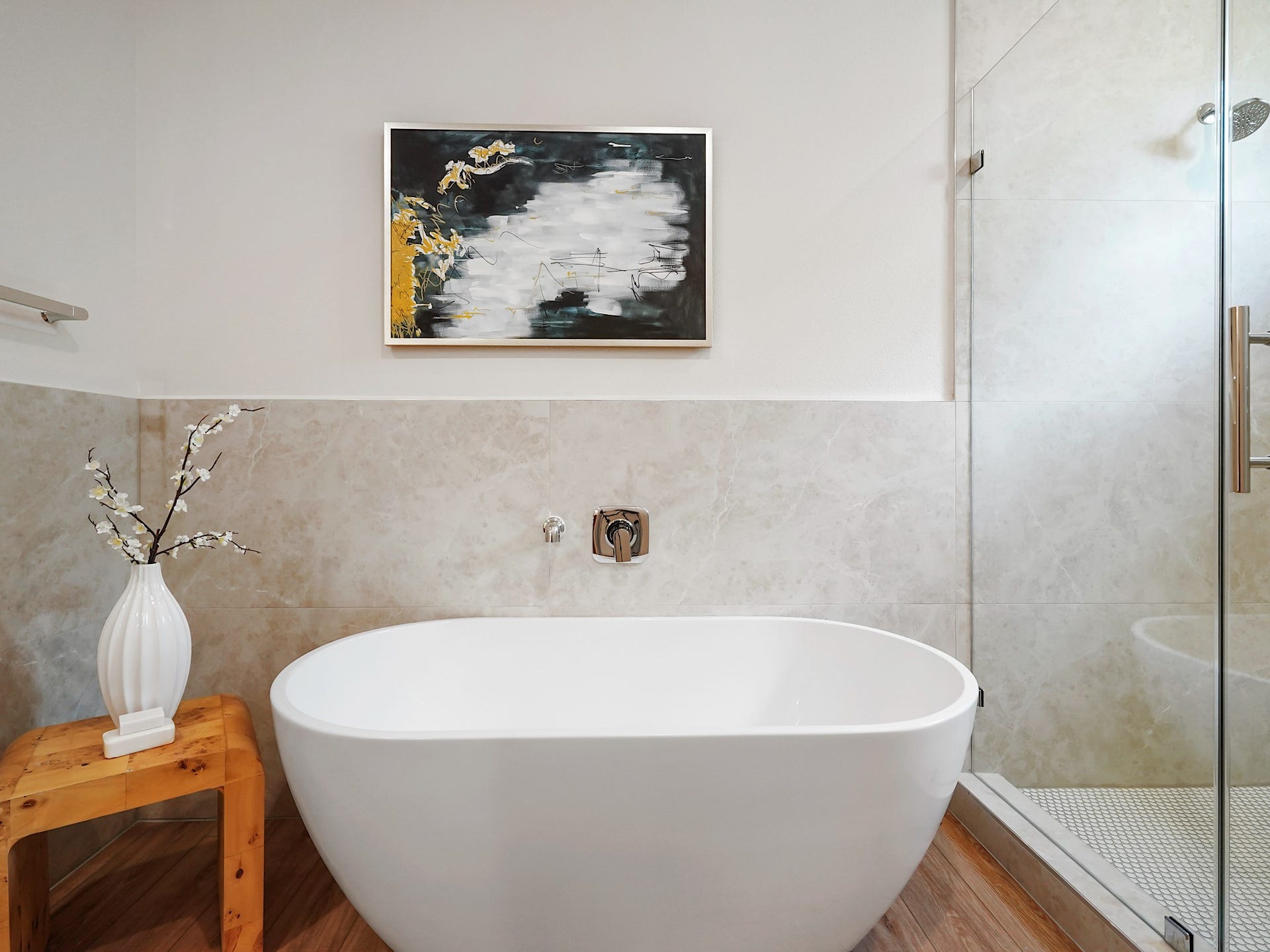 This bathroom features a modern freestanding tub, a glass-enclosed shower, and elegant tile work. A wooden stool with a vase adds warmth, while a contemporary artwork enhances the space's stylish appeal.