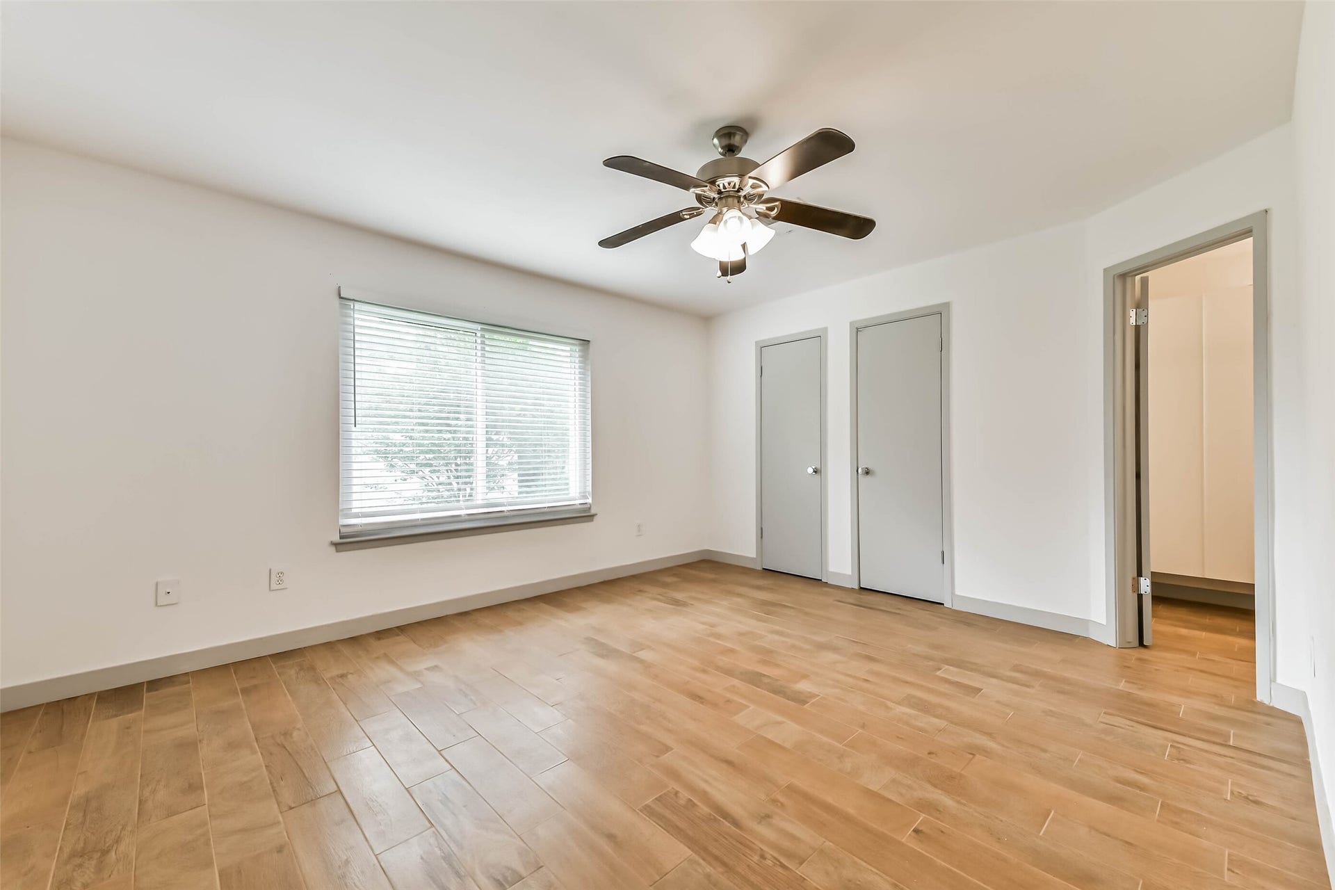 Bedroom features wood-look flooring, a ceiling fan, and ample natural light from a large window. It includes two closet doors and an additional doorway leading to another space.
