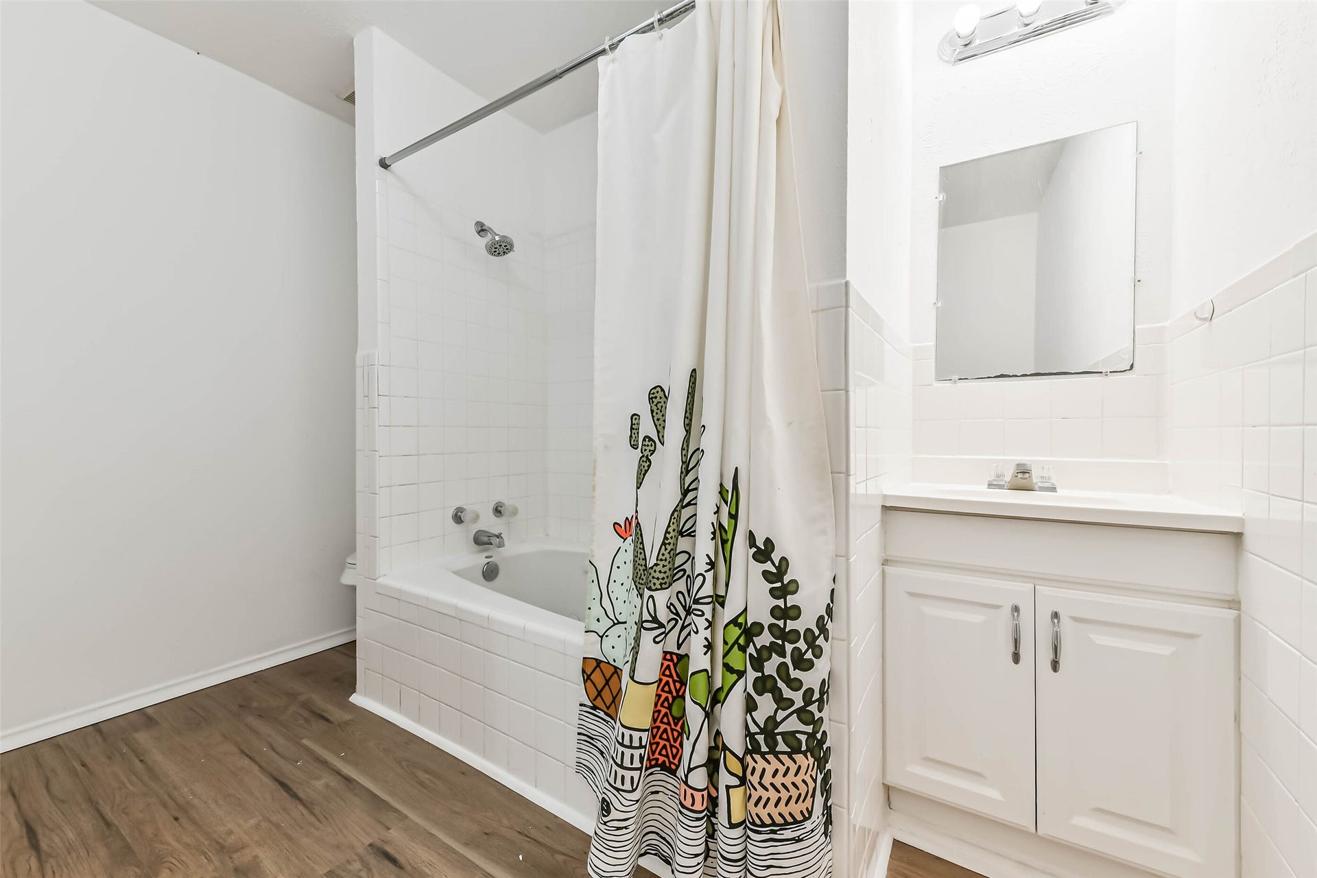 Bathroom features a clean, white design with a tiled shower and tub. It includes a vanity with storage, a large mirror, and wood-style flooring for a warm touch.