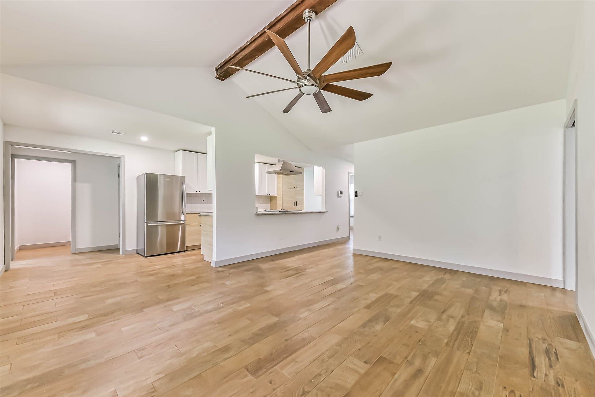 Open living space with light wood flooring and a ceiling fan. The room flows into a kitchen area with stainless steel appliances, including a refrigerator, and a partial view of white cabinetry.
