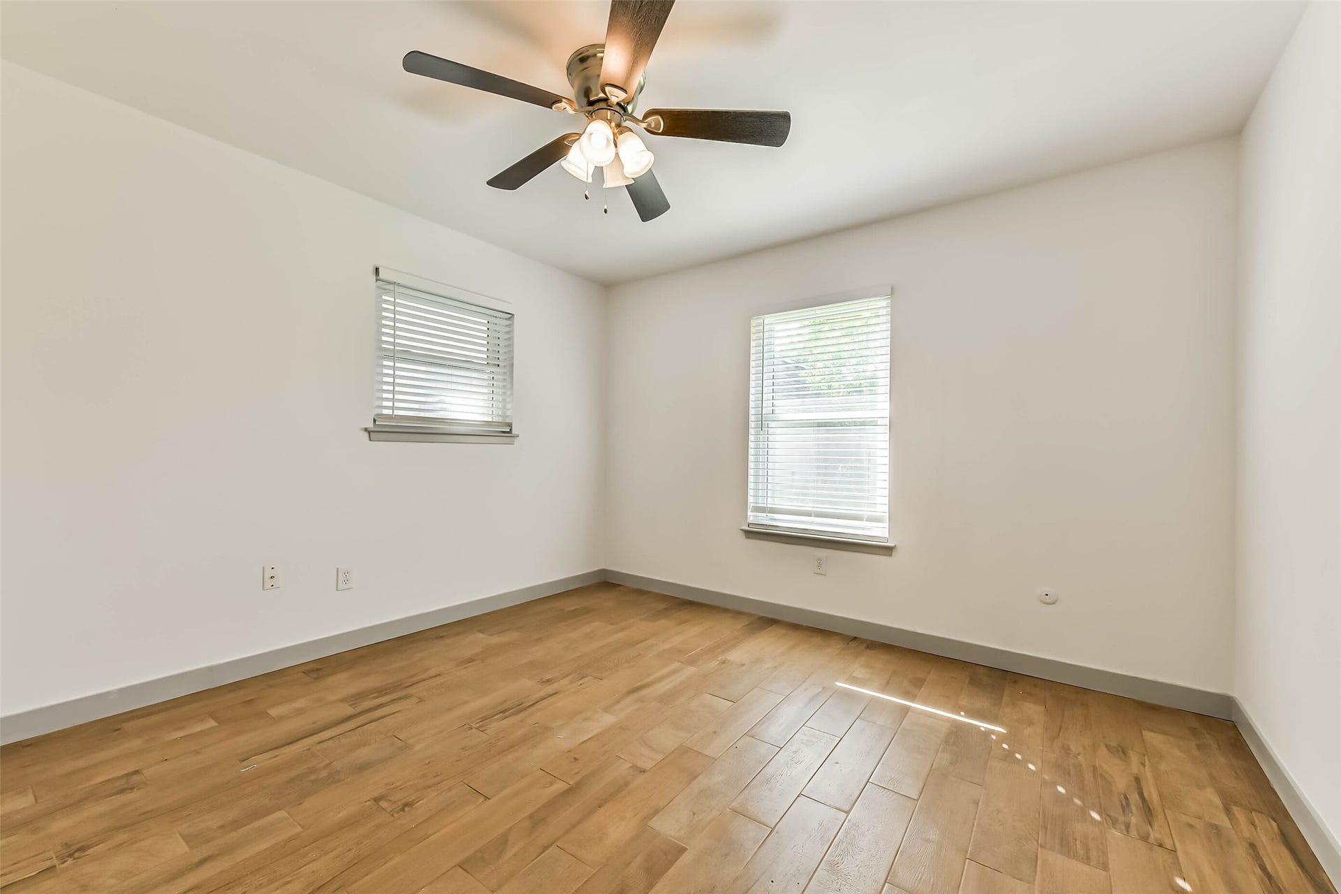 Bedroom features wood flooring, a ceiling fan, and two windows with blinds, offering a modern and clean look.