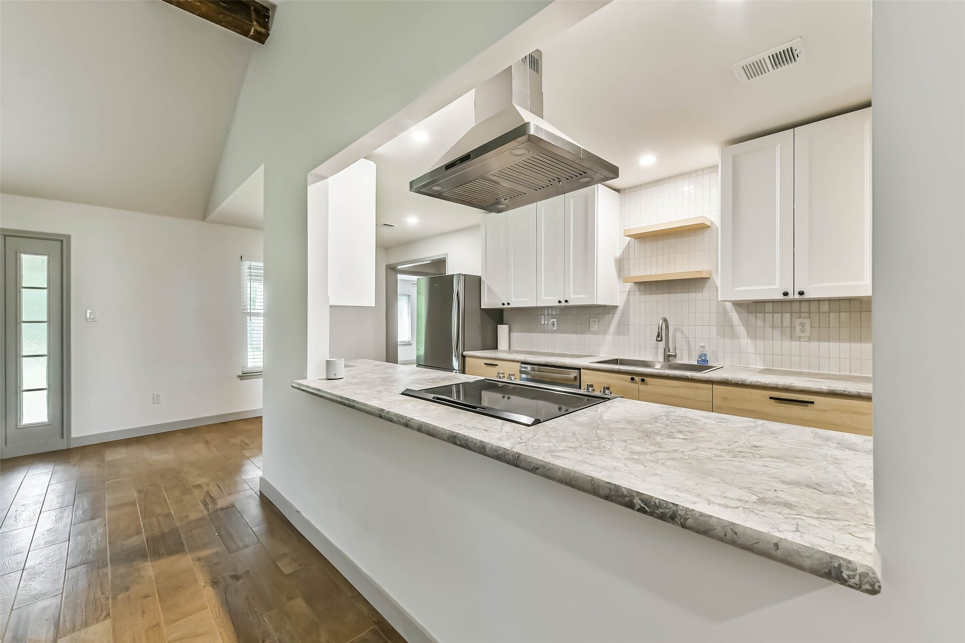 Modern kitchen with an open layout featuring marble countertops and a sleek range hood. The space includes white cabinetry, stainless steel appliances, and wooden flooring.