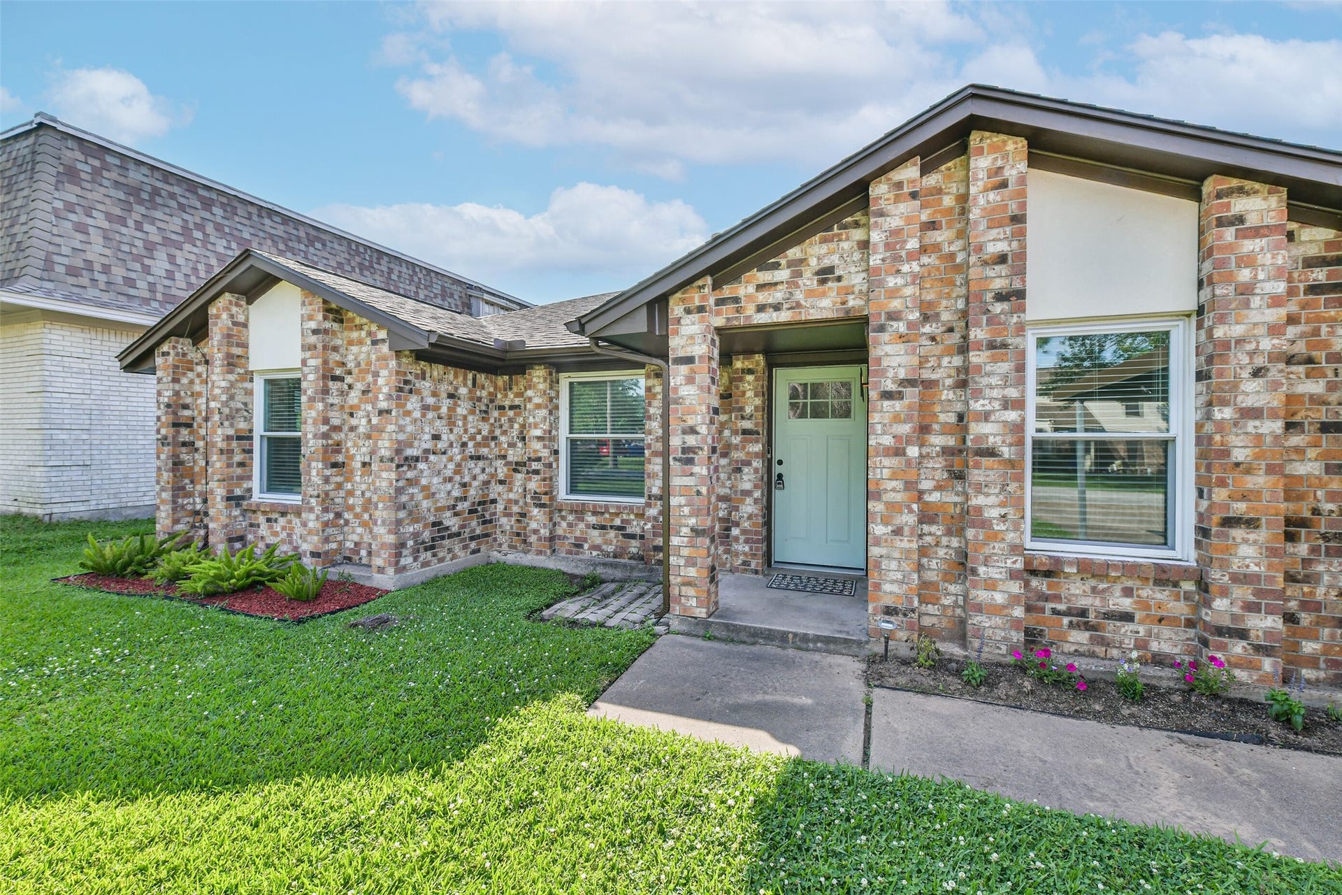 Charming brick home with a welcoming front porch and well-maintained landscaping. Features a modern front door, large windows, and a spacious lawn.