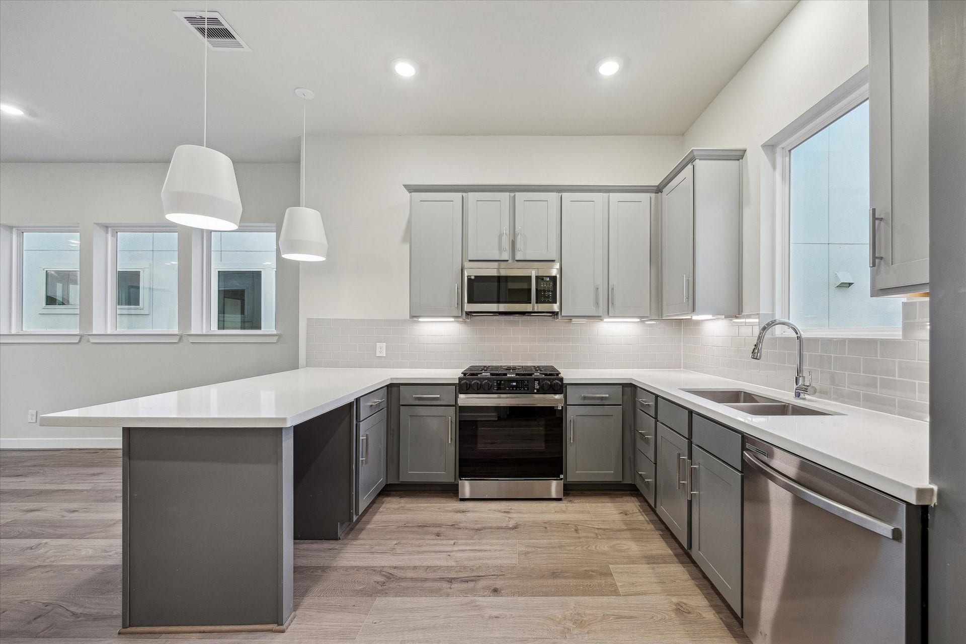 Kitchen featuring a large peninsula, gray cabinetry, white countertops, subway tile backsplash, pendant lighting, stainless steel appliances, and an open connection to the main living area.