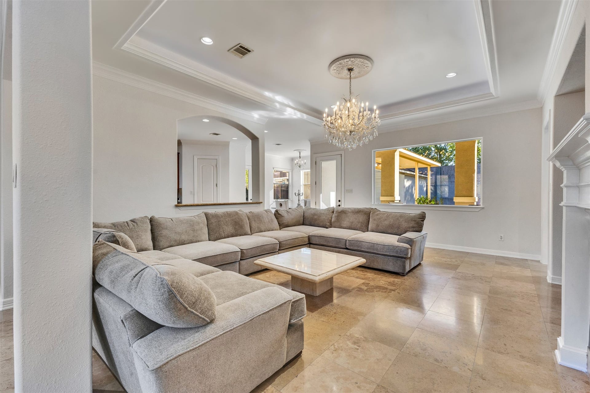 8606 Argentina Street, Houston, TX 77040 Elegant living room featuring high tray ceilings framed by intricate crown molding, a stunning chandelier with a decorative ceiling medallion, and graceful arched openings. Abundant natural light enhances the spacious, inviting atmosphere.