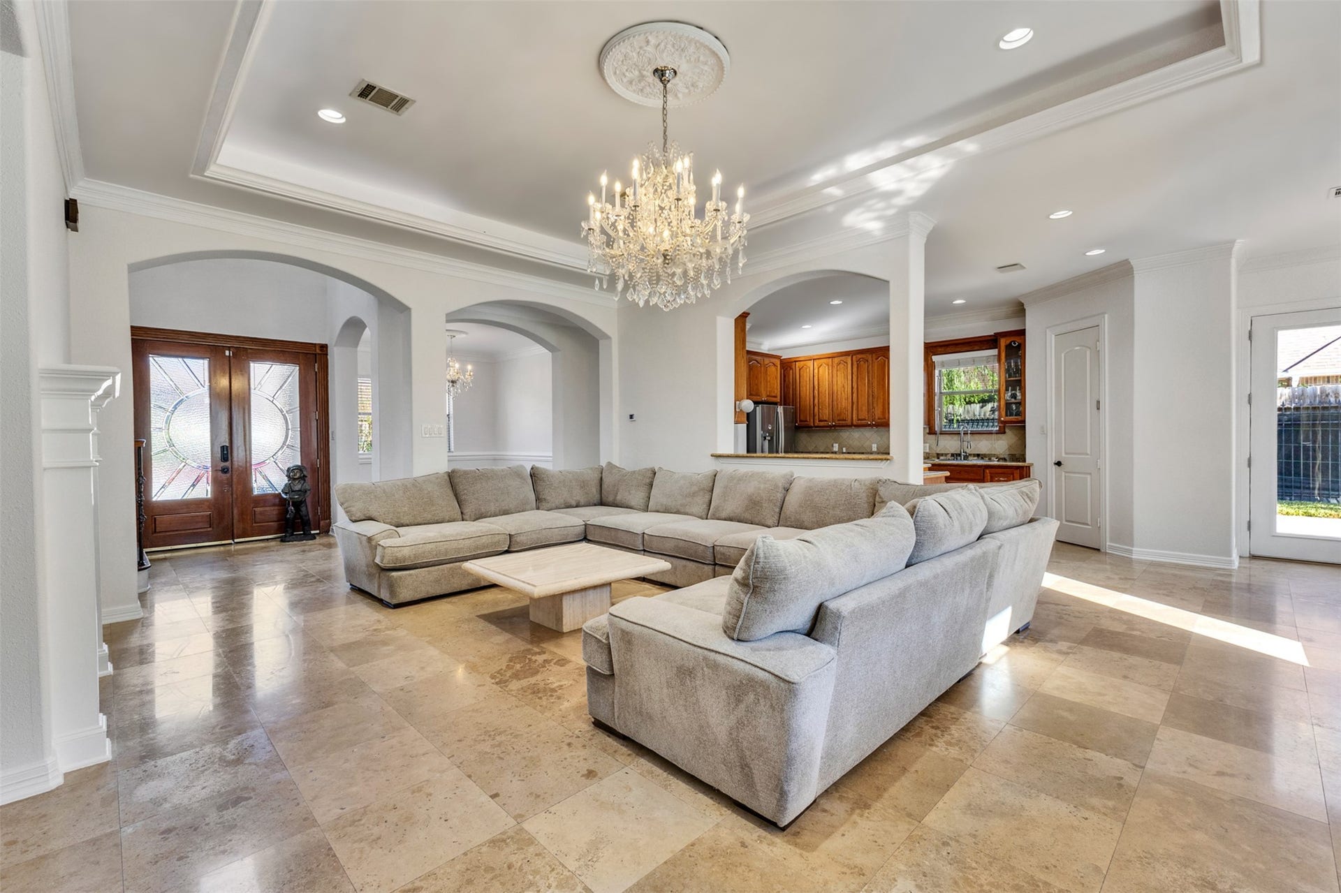 8606 Argentina Street, Houston, TX 77040 Elegant living room featuring high tray ceilings framed by intricate crown molding, a stunning chandelier with a decorative ceiling medallion, and graceful arched openings. Abundant natural light enhances the spacious, inviting atmosphere.