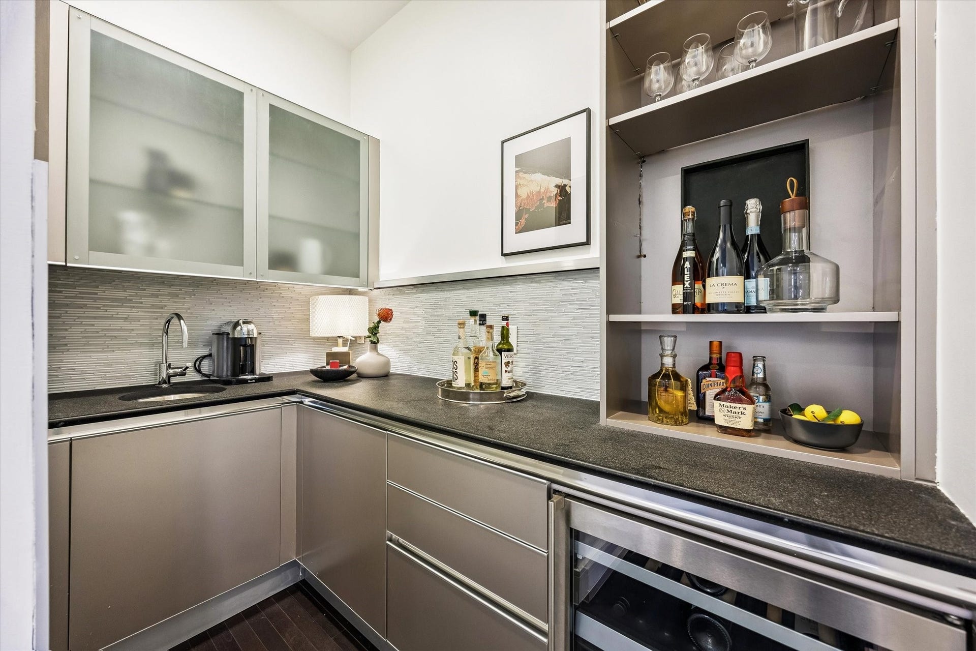 [Wet Bar] Wet bar features under cabinet lighting, glass display shelves, sink and wine refrigerator.