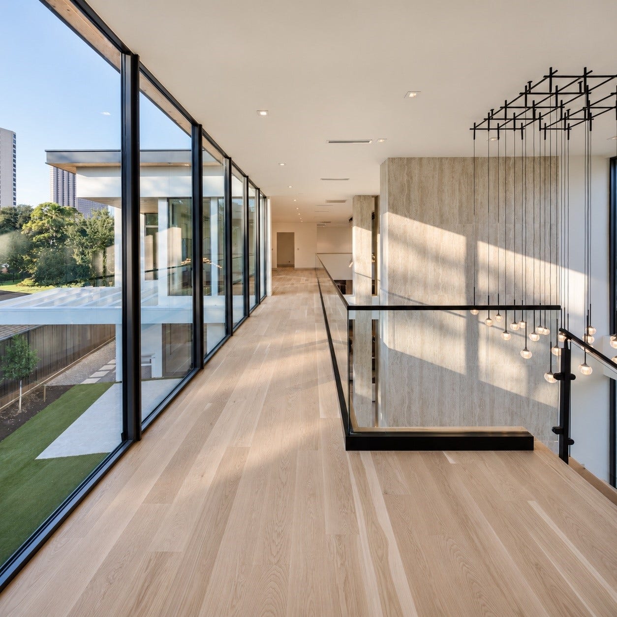 The second floor landing feels more like an elevate gallery in the sky. Wrapped in expansive floor-to-ceiling glass that brings the outdoors in. Glass railings presere the uninterrupted sightlines below, while warm wood floors and cascading light installation add softnerss and artistry