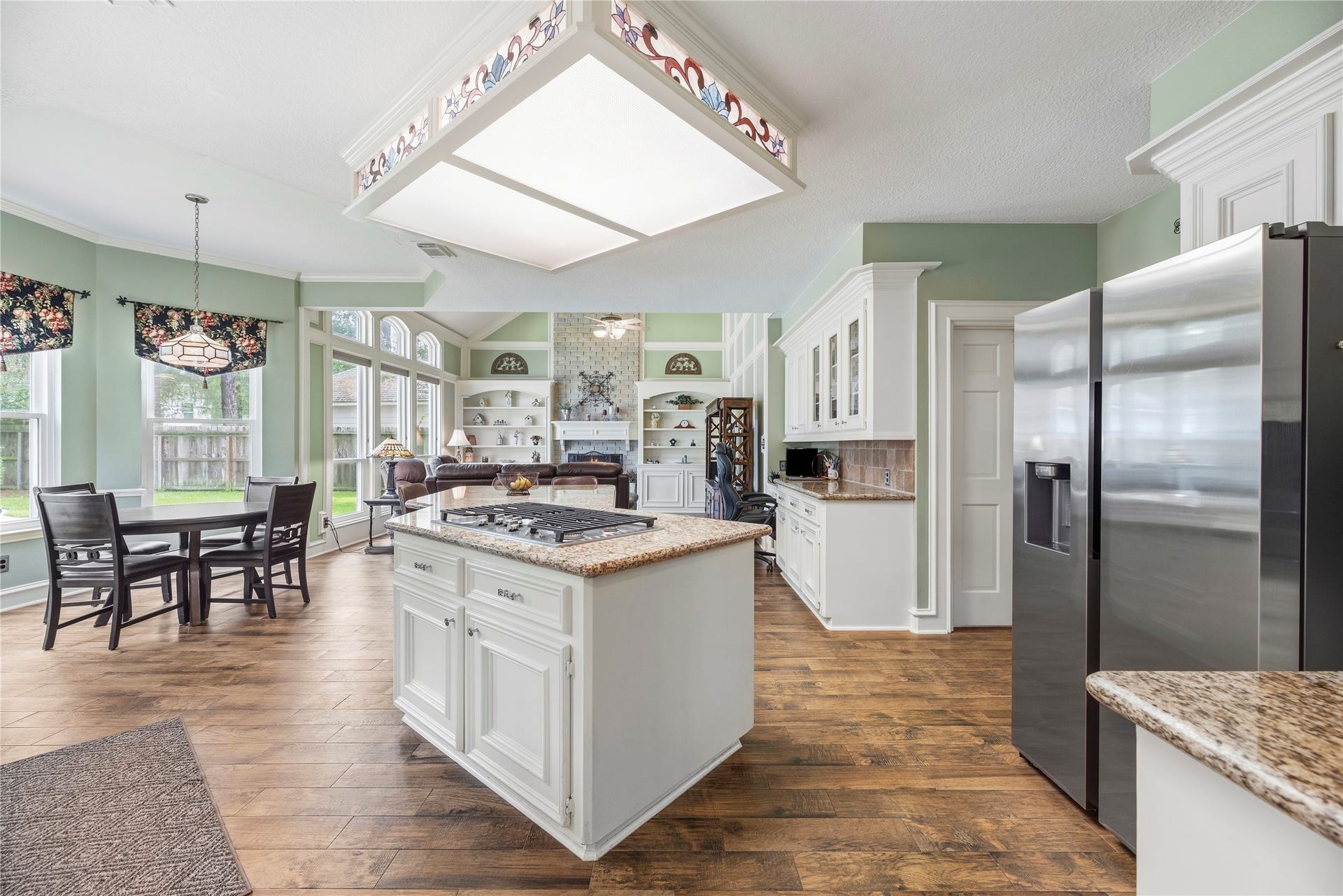 13114 S Winesap Lane, Cypress, TX 77429 Spacious open-plan kitchen and aeating area with elegant white cabinetry, granite countertops, and a central island cooktop. Abundant natural light flows through large windows, enhancing the warm wood flooring and creating a welcoming atmosphere.