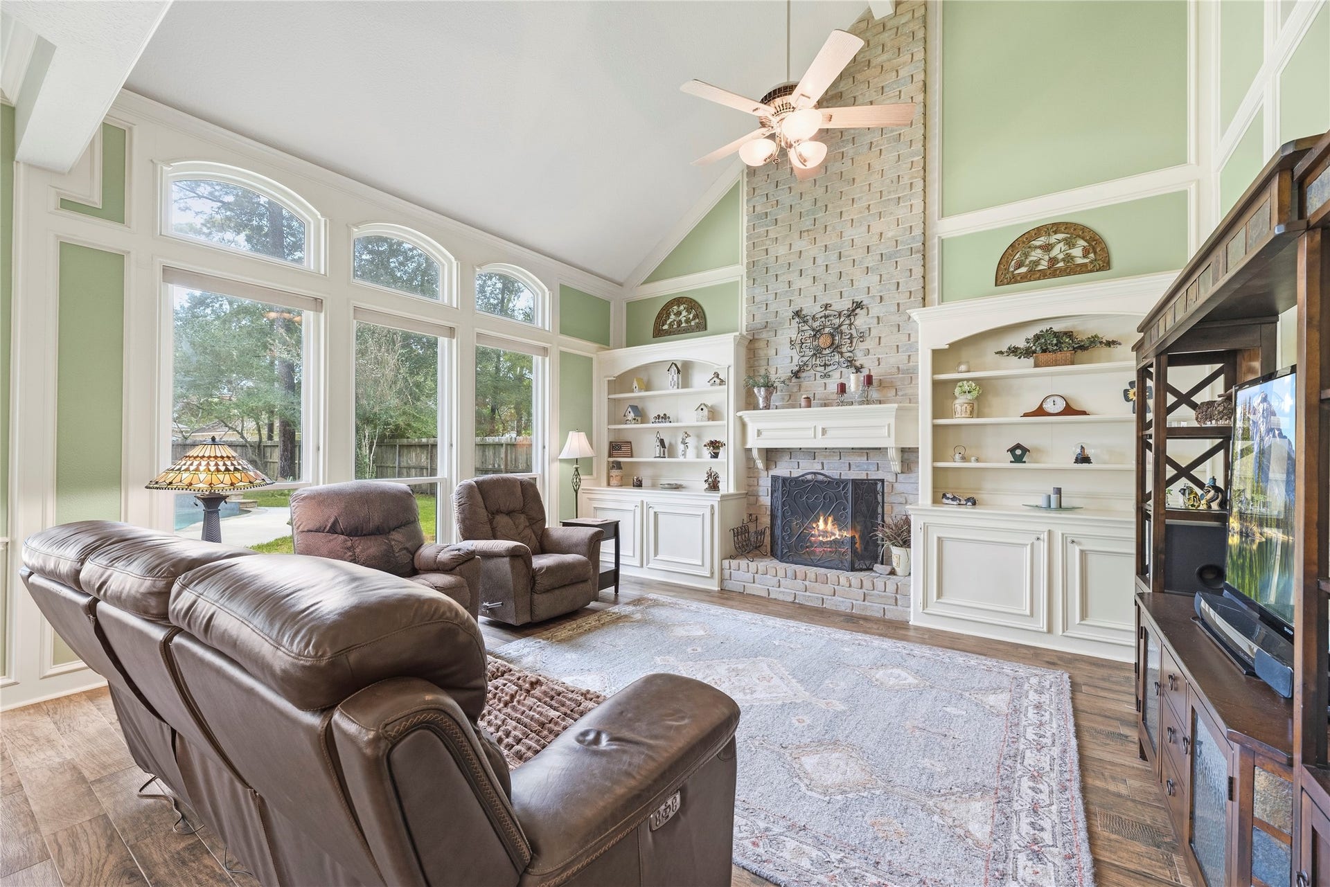 13114 S Winesap Lane, Cypress, TX 77429 Spacious living room with high vaulted ceilings, featuring a cozy fireplace, built-in shelves, and large arched windows offering a serene view of the outdoors that flood the room with natural light.