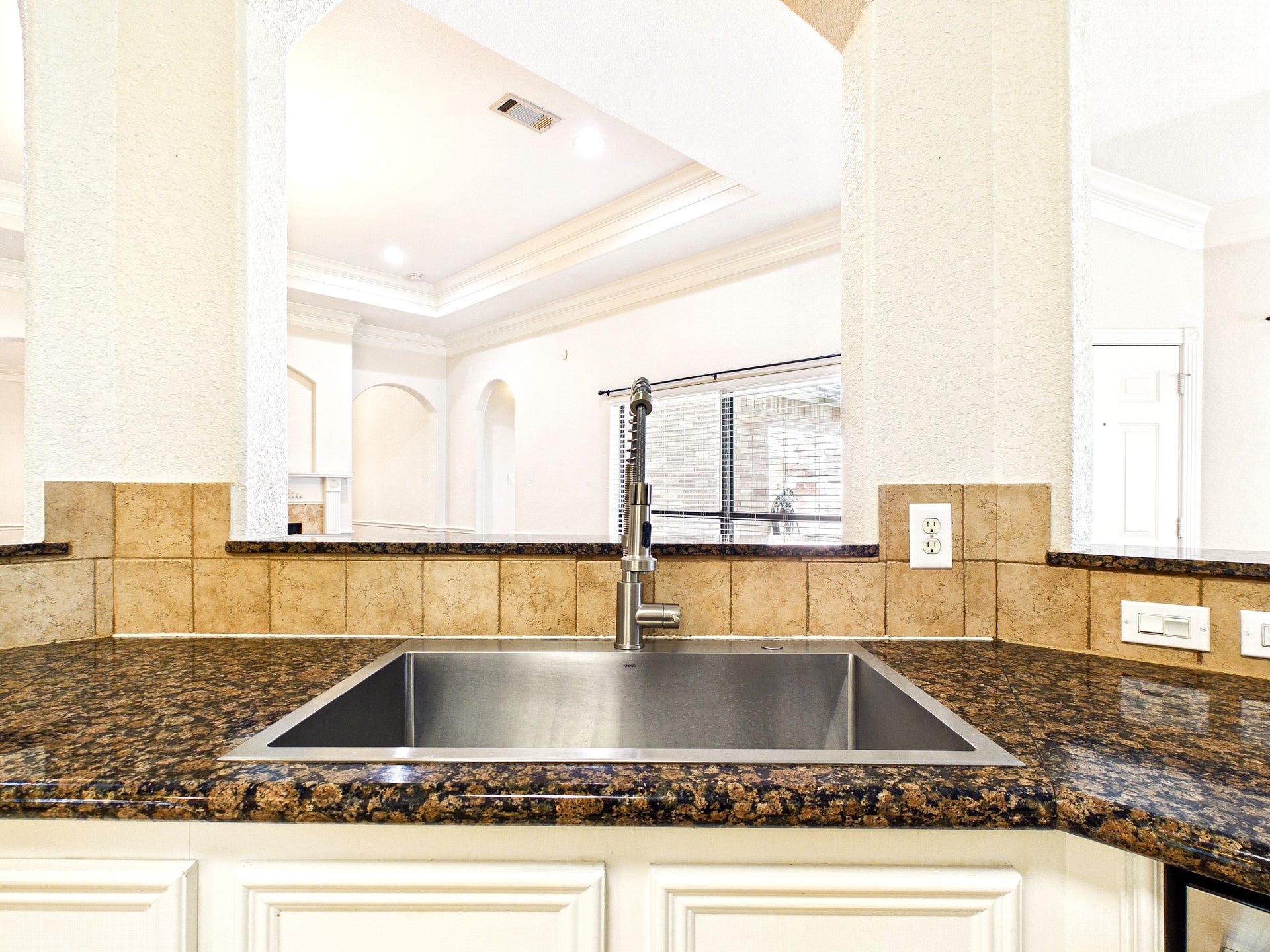 19521 Desna Drive, Porter, TX 77365 Modern kitchen area with a stainless steel sink, granite countertops, and tile backsplash. The open layout offers a view into a bright living space with crown molding and large windows.