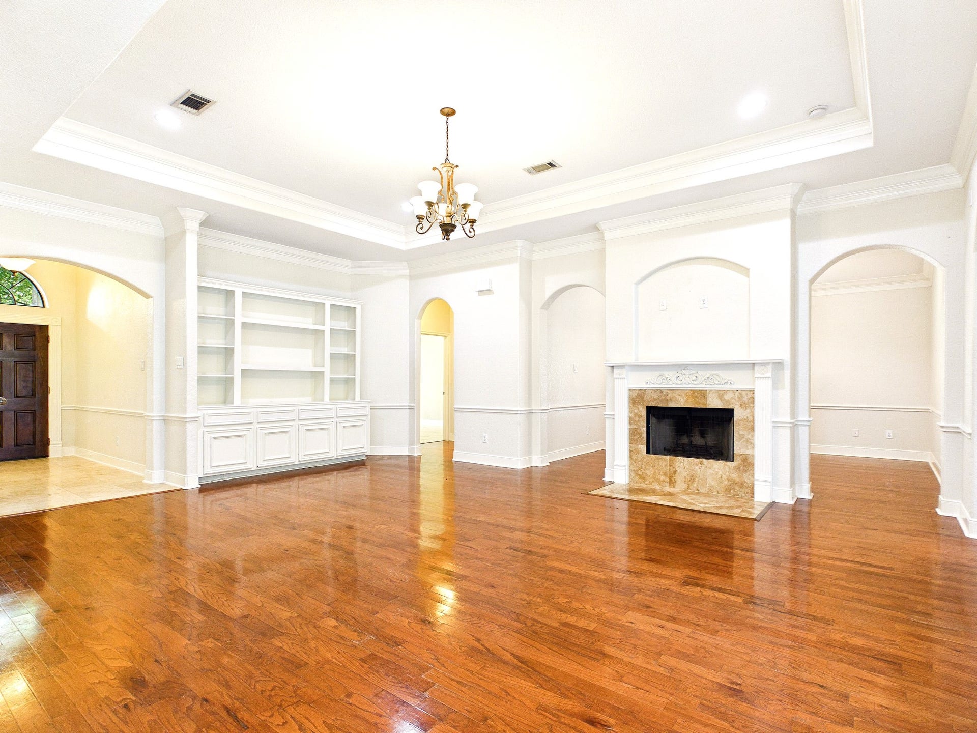 19521 Desna Drive, Porter, TX 77365 This spacious living room features elegant hardwood floors, a decorative fireplace, built-in shelving, and a tray ceiling with a classic chandelier. Arched doorways add a touch of sophistication, while abundant natural light enhances the warm ambiance.