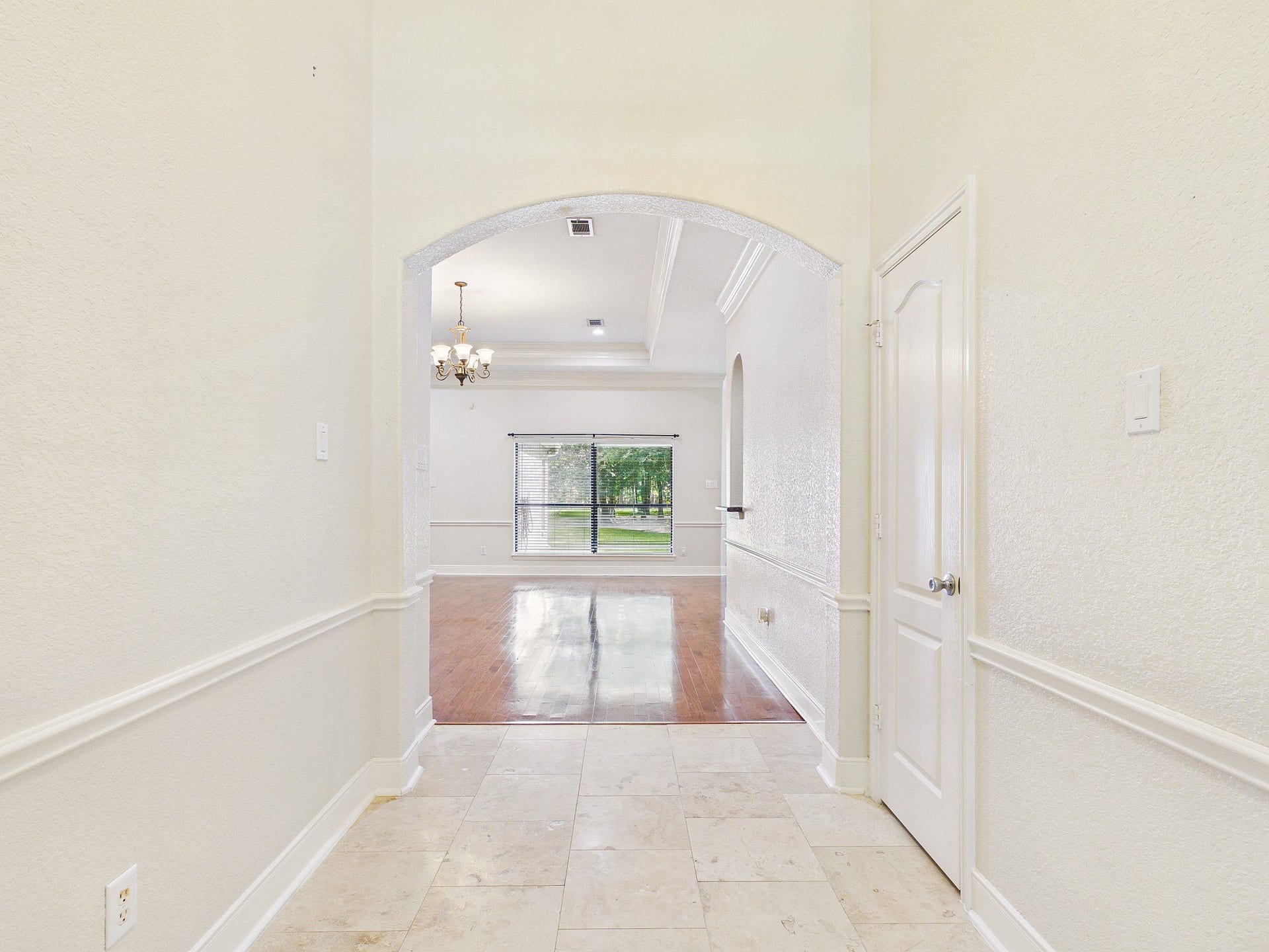 19521 Desna Drive, Porter, TX 77365 The photo shows a bright, open hallway leading into a spacious living area. The hallway features tiled flooring, while the living area has wooden floors. A large window at the far end provides ample natural light, and a chandelier adds elegance to the space.
