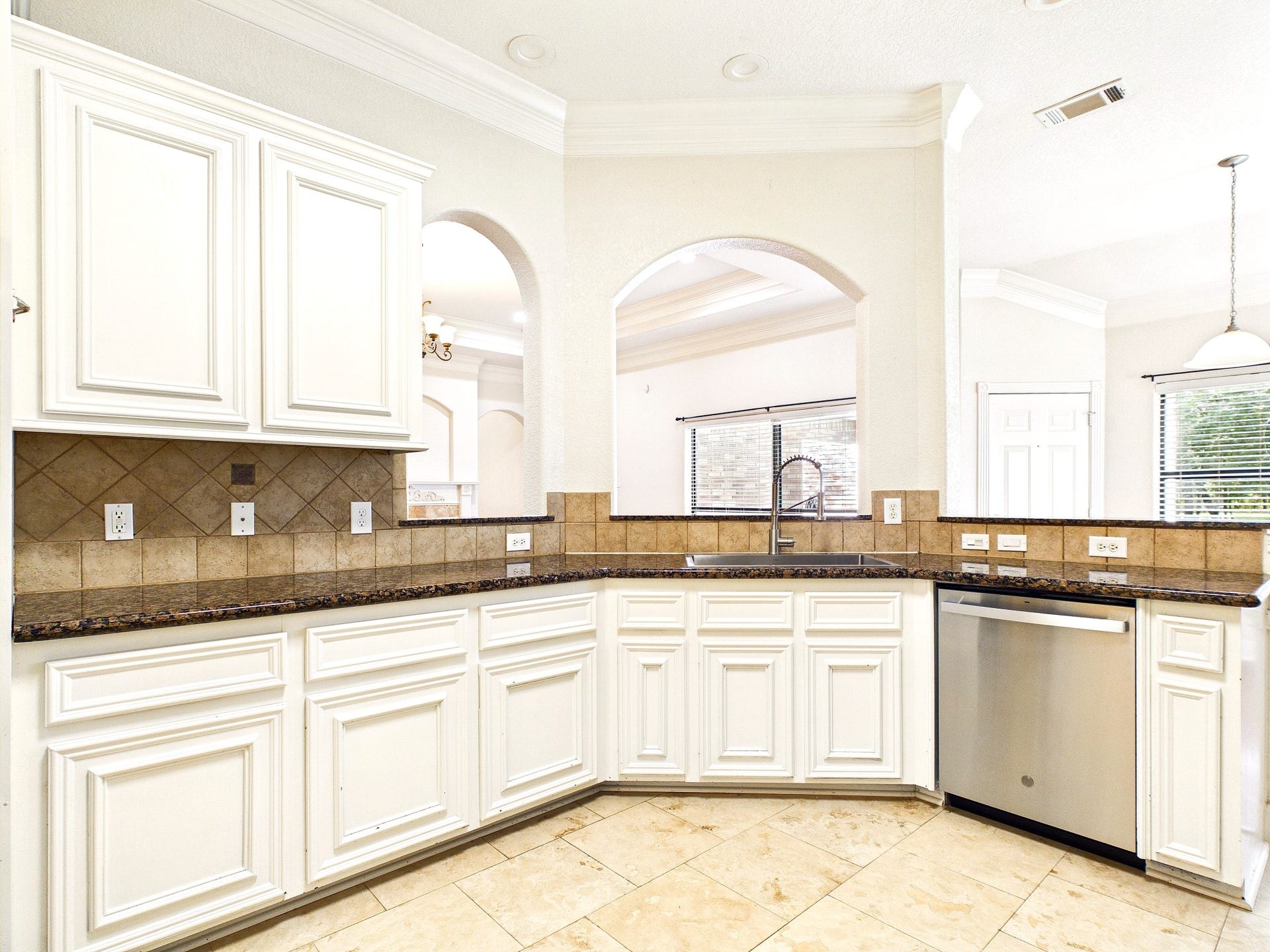 19521 Desna Drive, Porter, TX 77365 This kitchen features elegant white cabinetry, granite countertops, and a stainless steel dishwasher. It has a tile backsplash and floor, with large windows offering plenty of natural light. The open layout includes arched openings connecting to adjacent rooms.